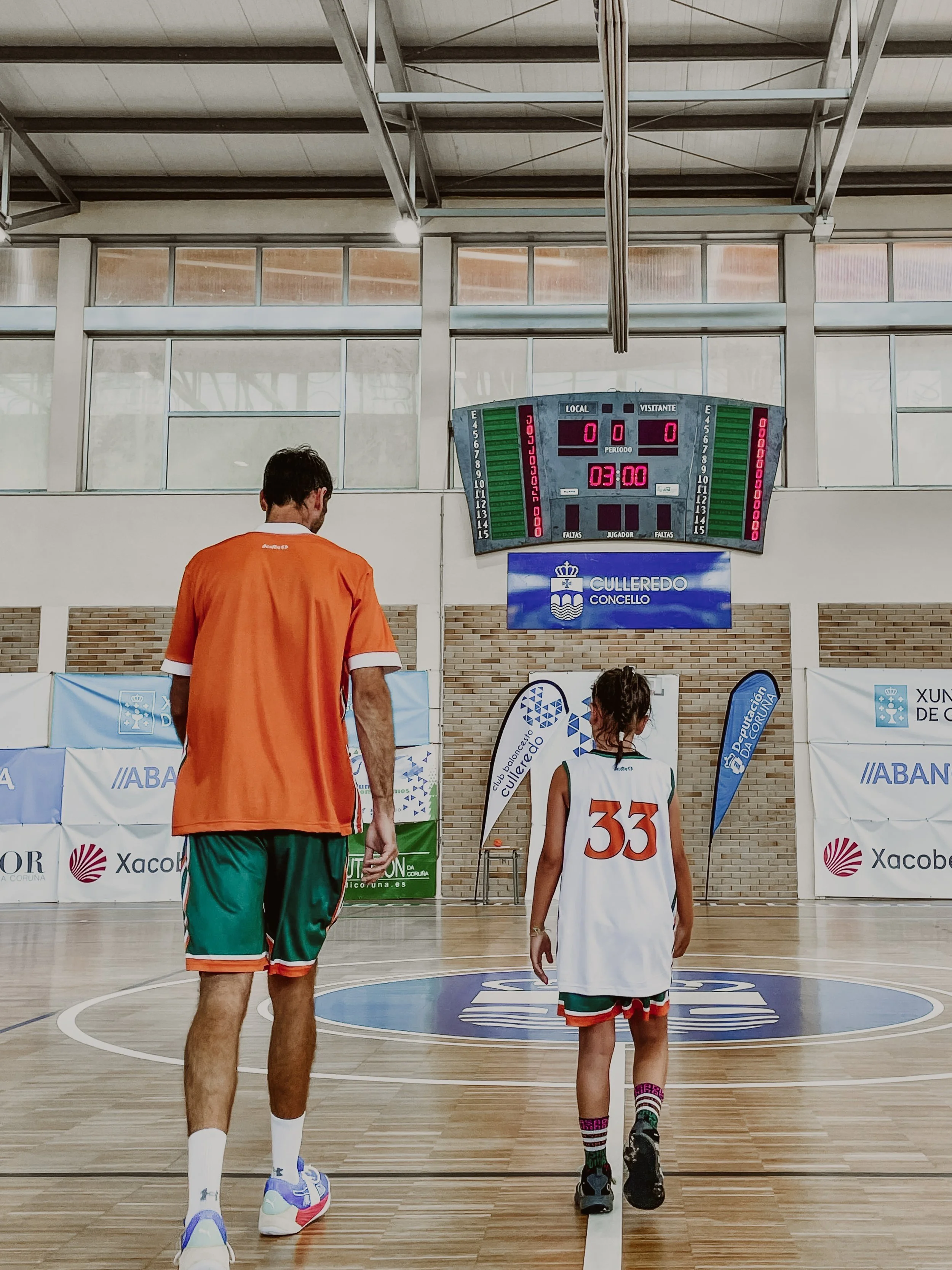Un entrenador y una joven jugadora de baloncesto caminando en una cancha de interior, con marcador digital y banderas de fondo.