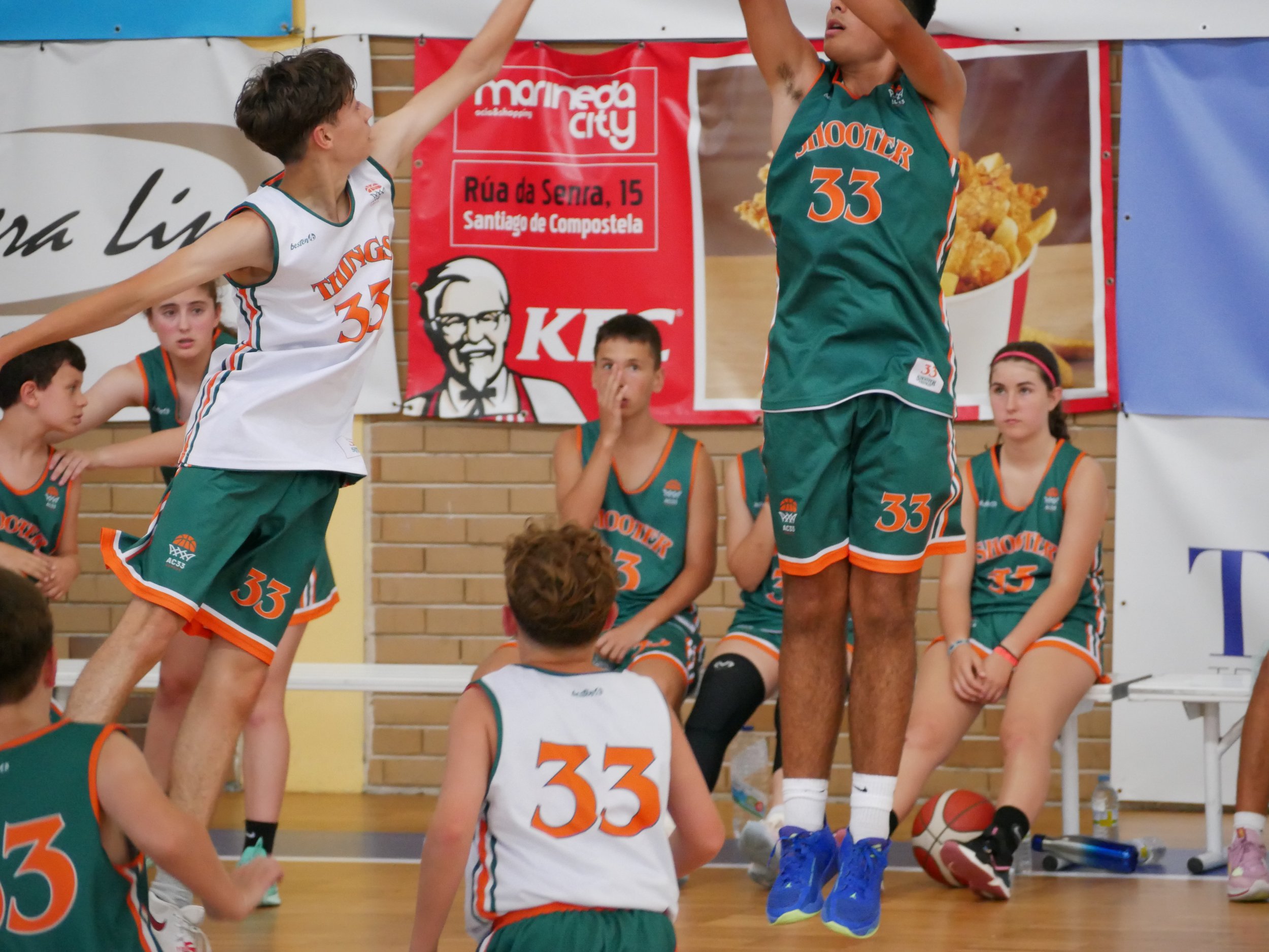 Jugadores de baloncesto niños en un juego, con un equipo de camisetas blancas y verdes con naranja, uno saltando para bloquear una tira, mientras otros observan y están sentados o de pie en el fondo.
