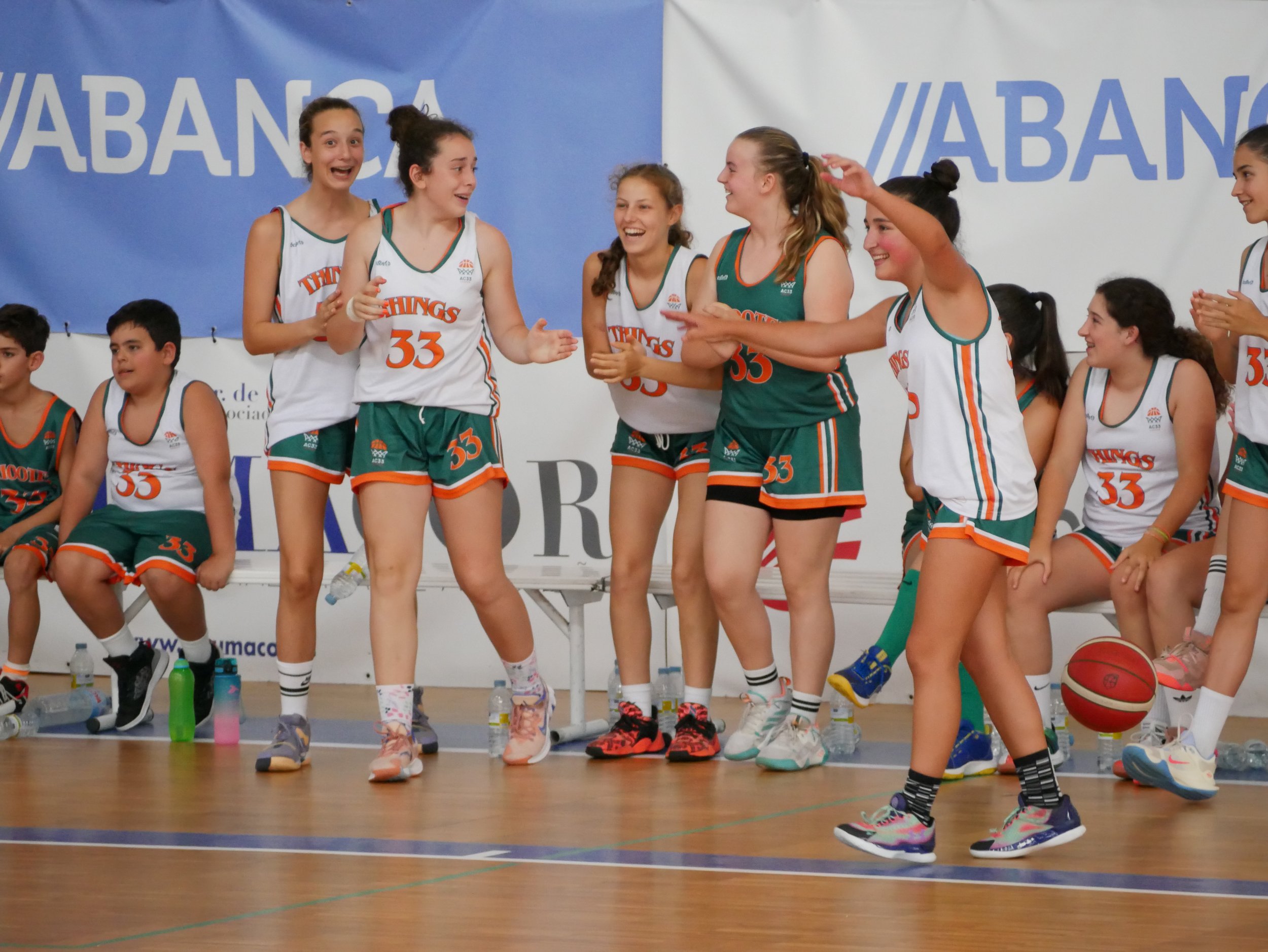 Niñas jugando baloncesto en un equipo, algunas están de pie y otras sentadas, en un ambiente de competencia deportiva