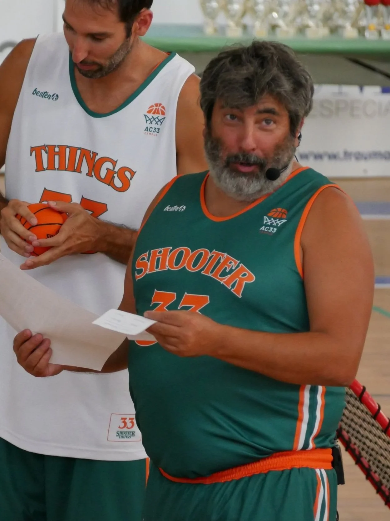 Dos hombres en un partido de baloncesto, uno con barba y cabello oscuro, vestido con uniforme verde con naranja que dice 'Shooter 33', y otro con barba canosa, con uniforme blanco que dice 'Things 57', en una cancha de baloncesto.