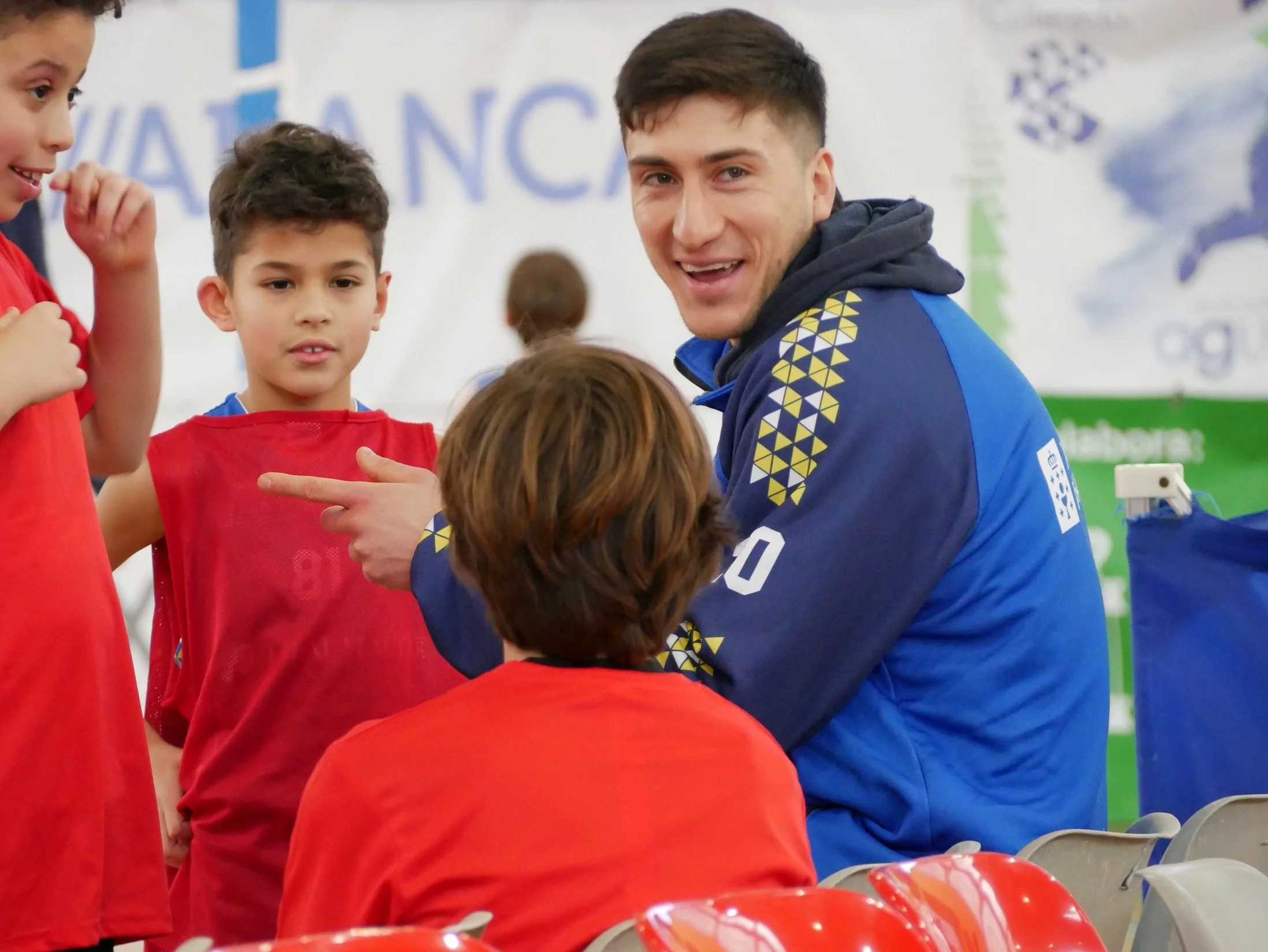 Un entrenador con camiseta azul y niños en uniformes deportivos rojos. El entrenador está hablando con un niño de cabello castaño y en el fondo hay una lona con texto y logotipos deportivos.
