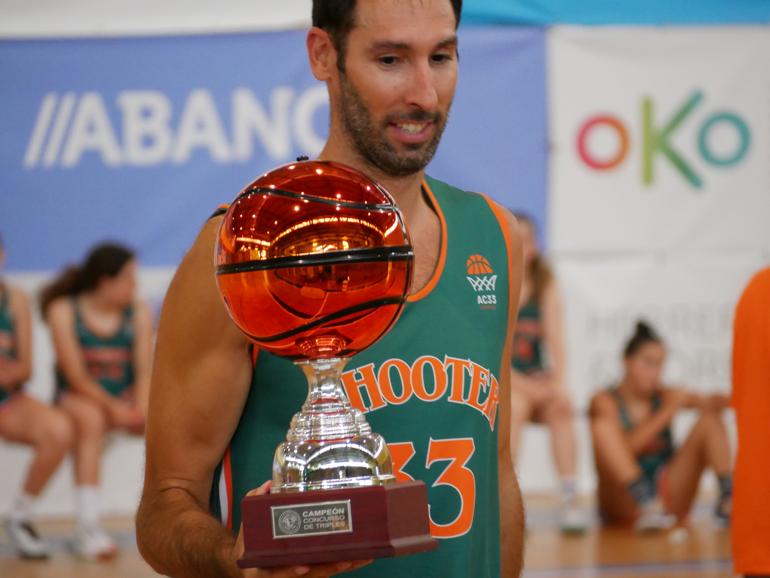 Jugador de baloncesto con uniforme que sostiene un trofeo con una pelota de baloncesto en la parte superior en un entorno de cancha.