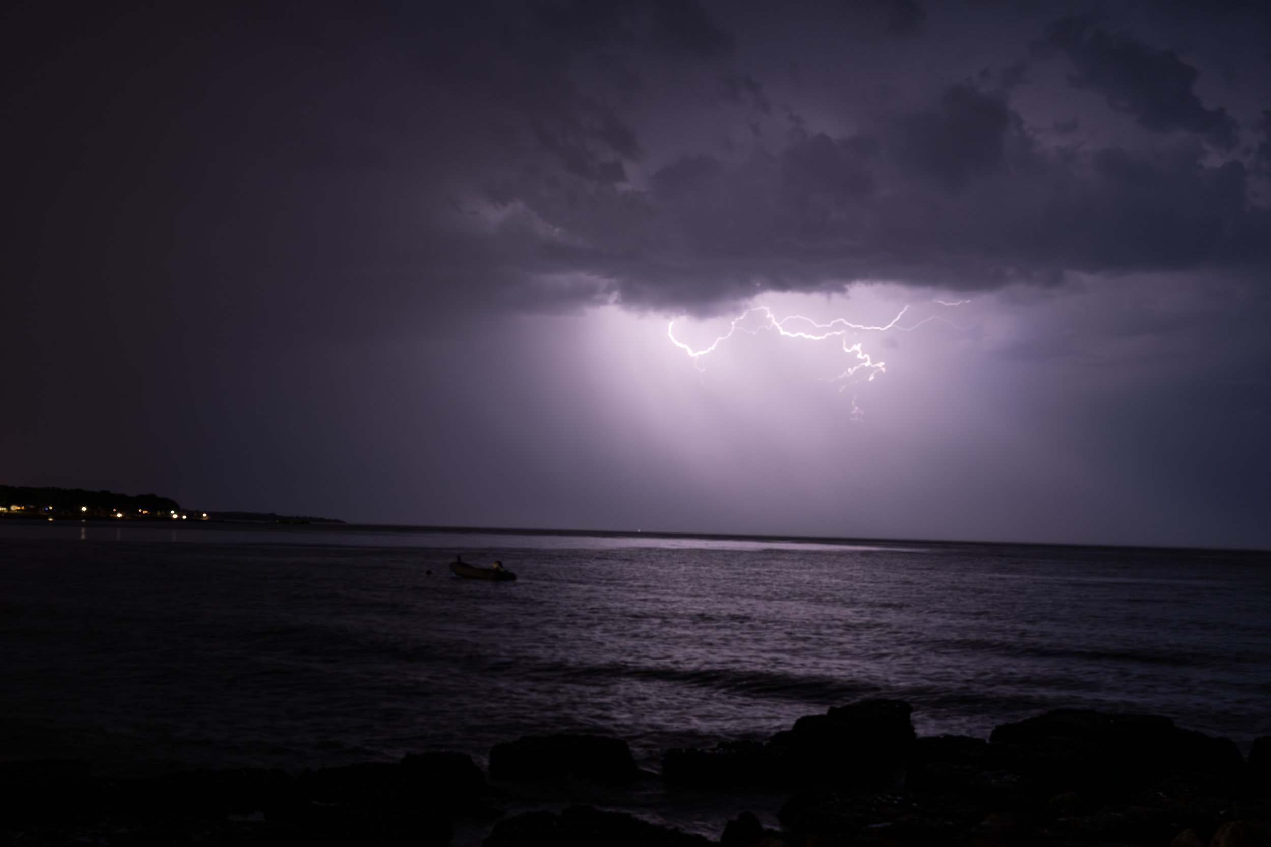 Stürmisches Gewitter mit Blitzen über dem Meer bei Nacht, dunkler Himmel, ferne Lichter am Horizont, Felsen im Vordergrund.