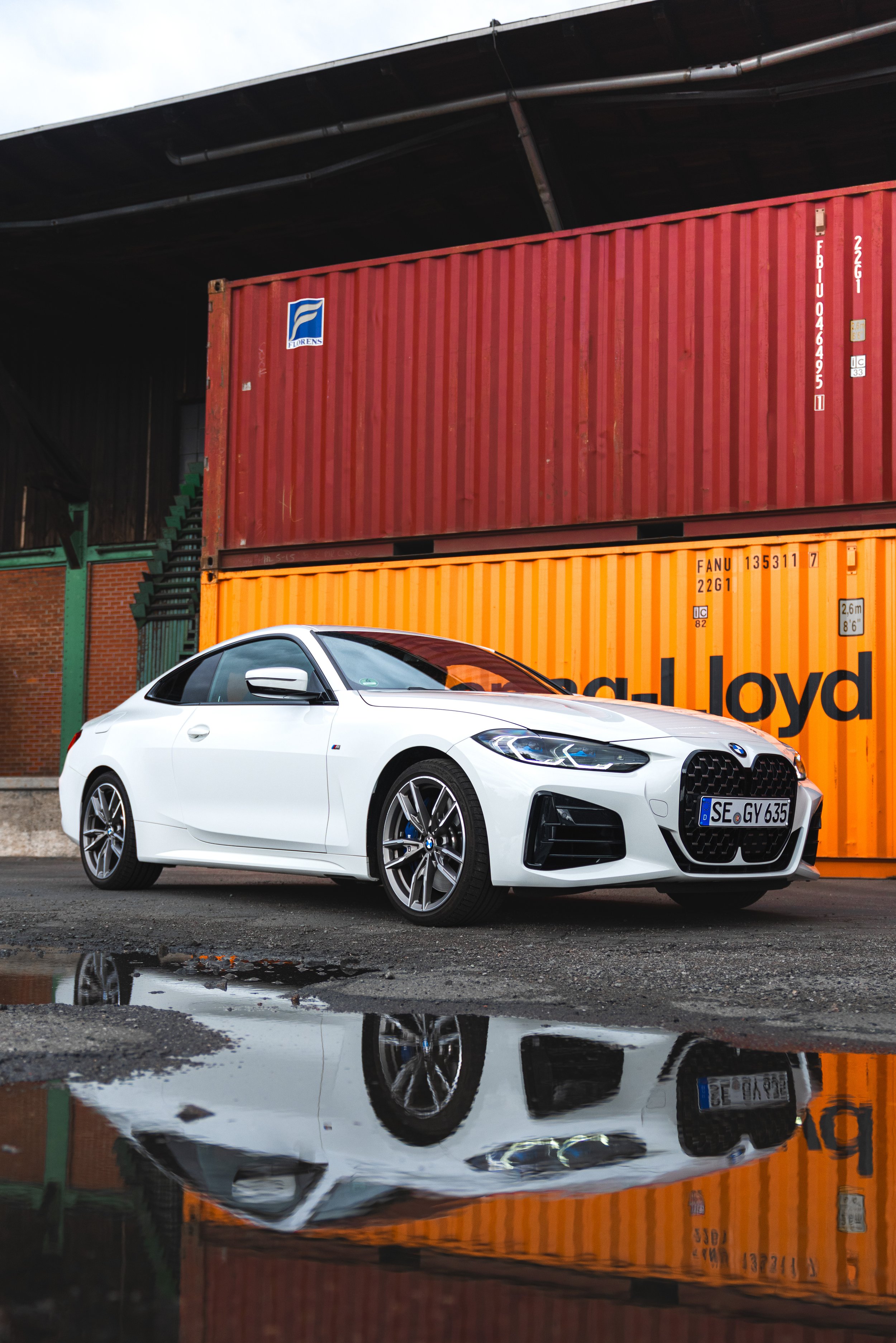 Weißer BMW-Coupé vor mehreren gelben und roten Container-Transportkästen auf einer nassen Straße, mit Reflexion im Wasser