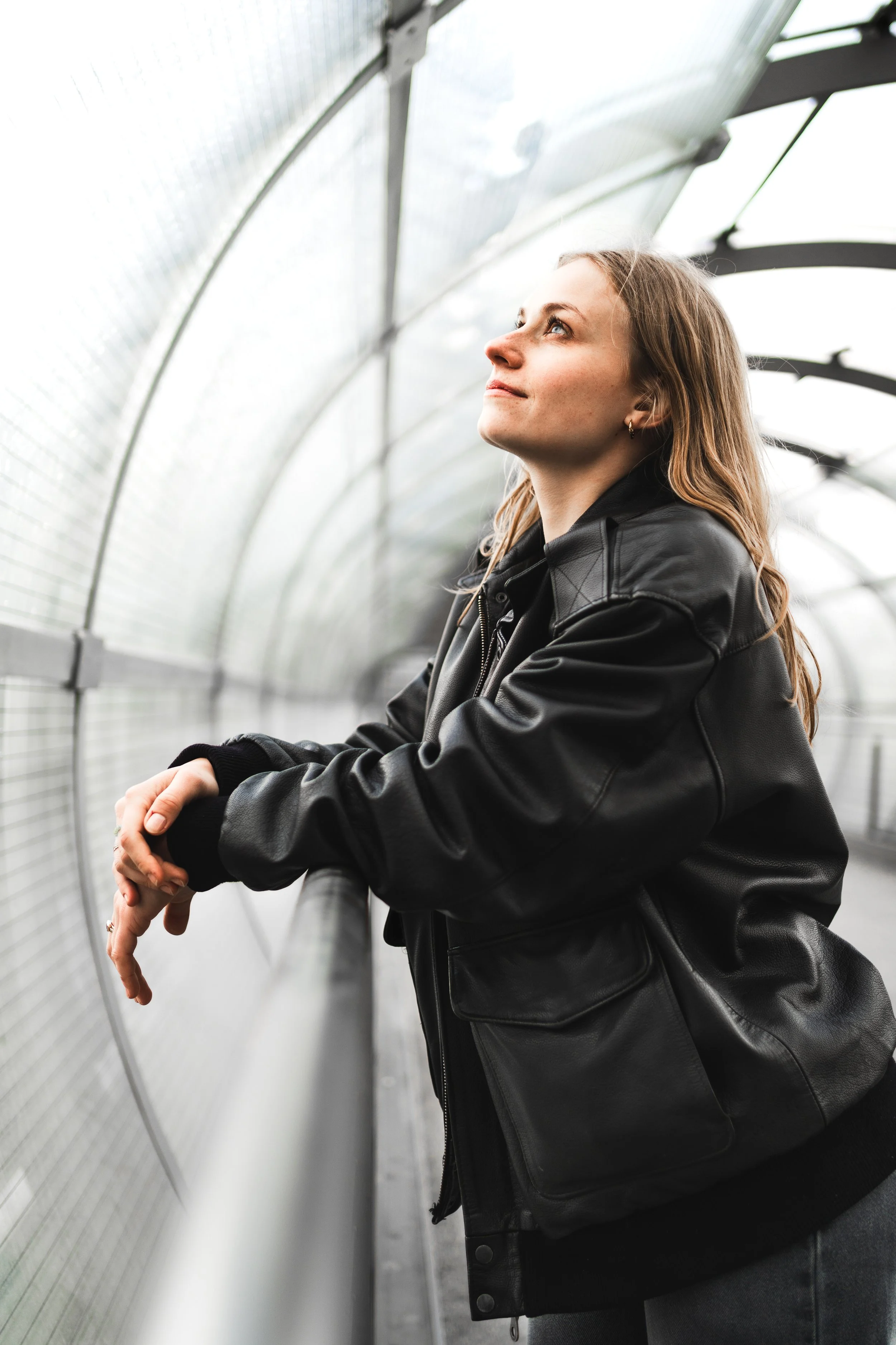 Eine junge Frau mit braunen Haaren, schwarze Lederjacke, sitzt in einem Gewächshaus und blickt nach oben.
