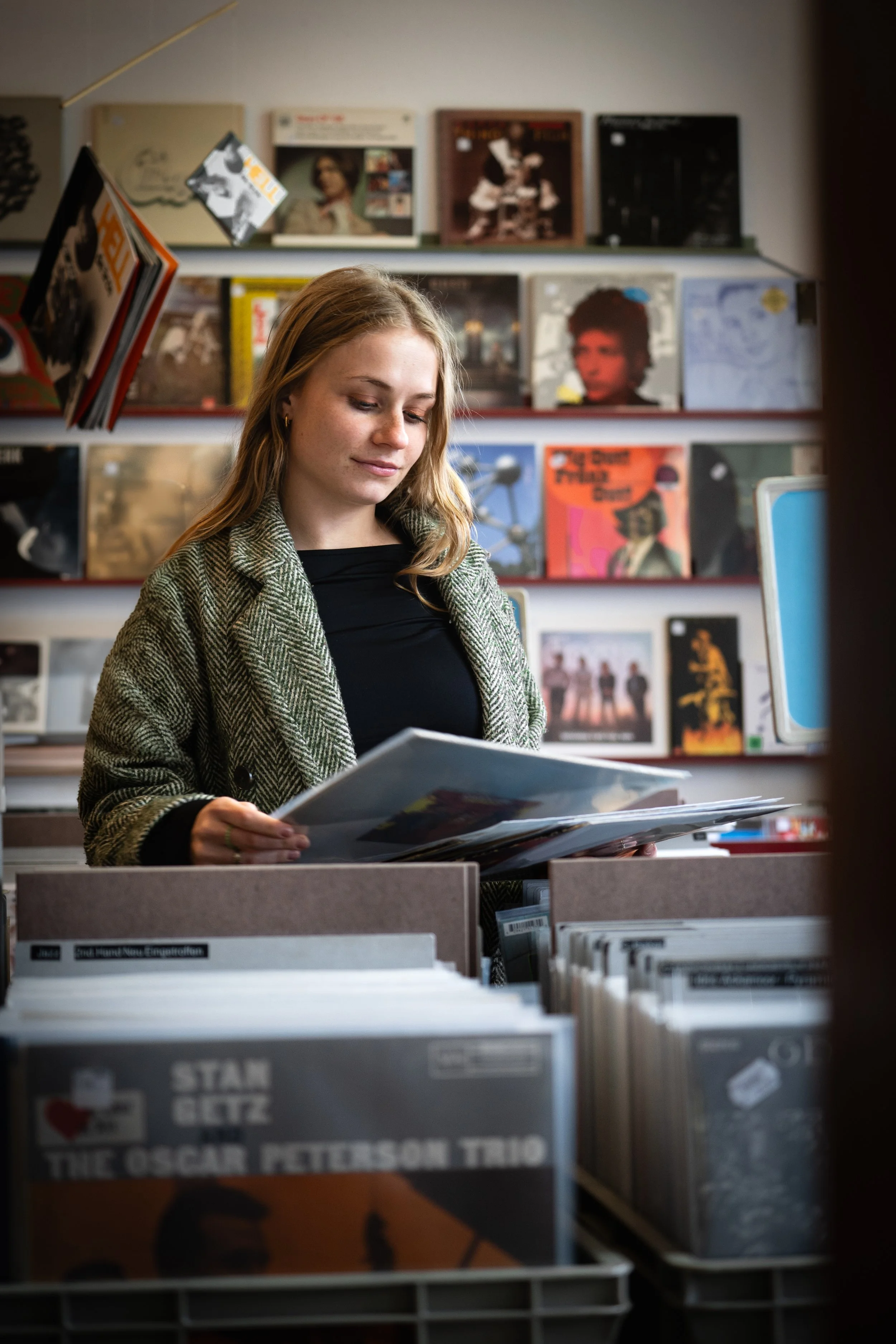 Eine junge Frau in einem Plattenladen, die in einem Album blättert, mit einer Sammlung von Vinyl-Schallplatten und einem Bücherregal im Hintergrund.