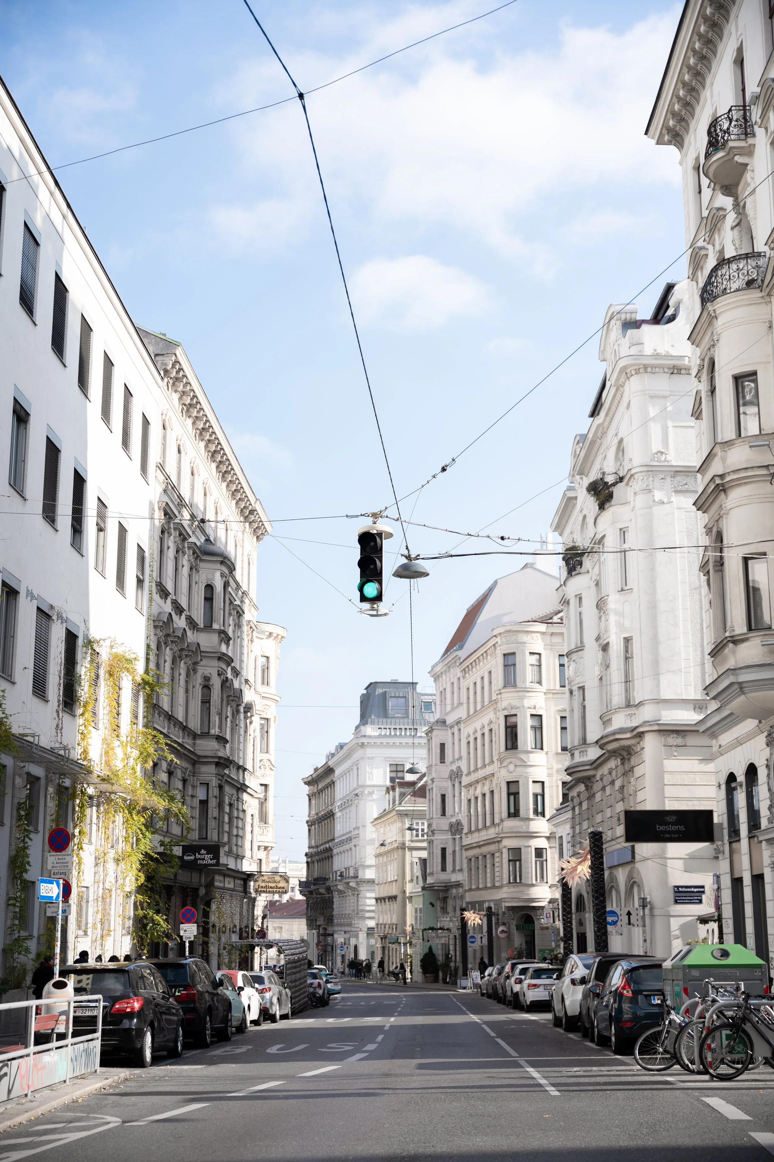 Eine Straße in einer europäischen Stadt mit weißen historischen Gebäuden, Autos parken an beiden Seiten, eine Ampel mit grünem Licht, und Drahtseilen für Tram- oder Busverkehr, während der Himmel blau mit wenigen Wolken ist.