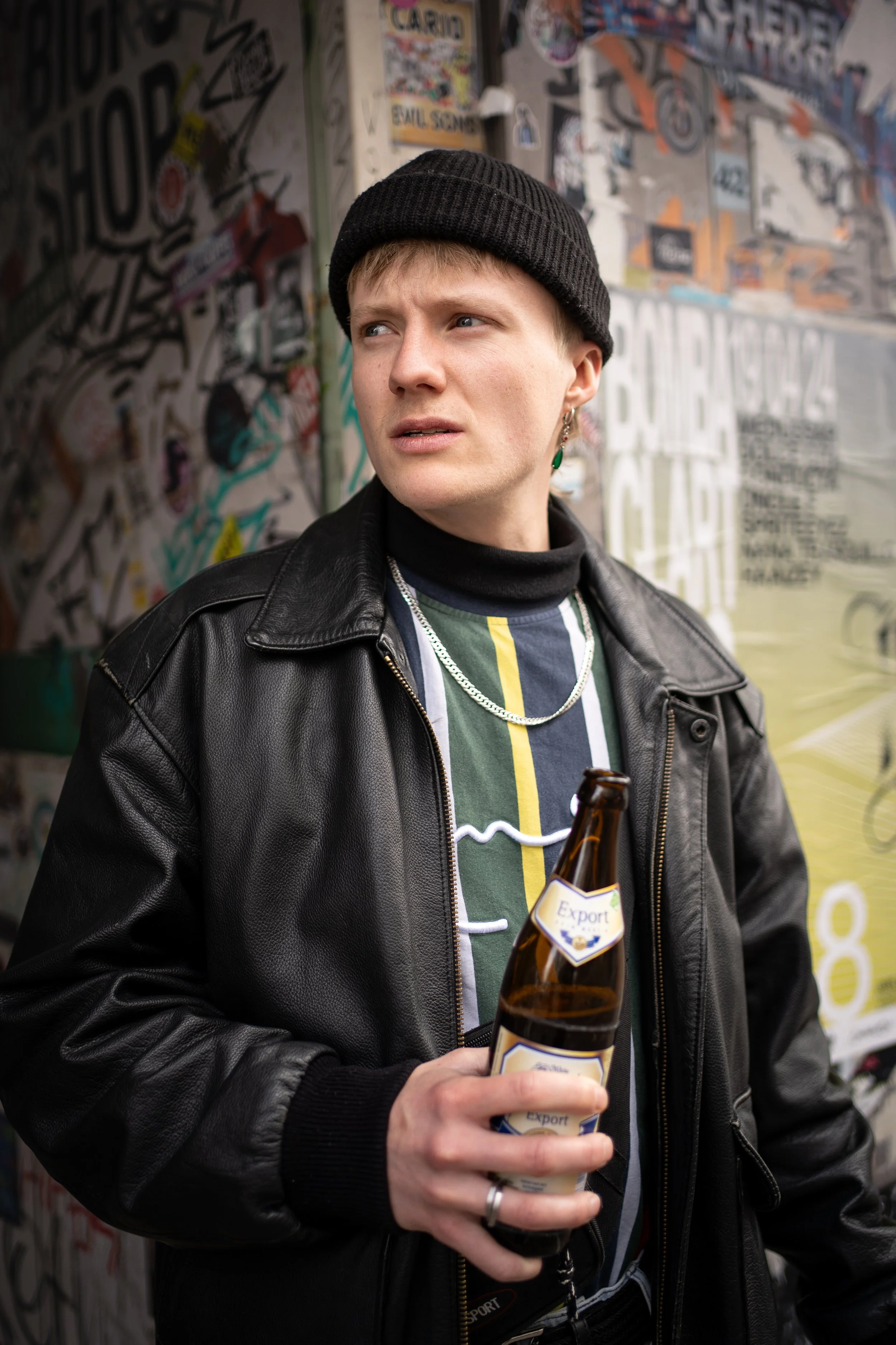 Junge Person mit schwarzen Ledermantel, schwarzen Mütze und Earring hält eine Bierflasche in der Hand vor einer Graffiti-Wand.