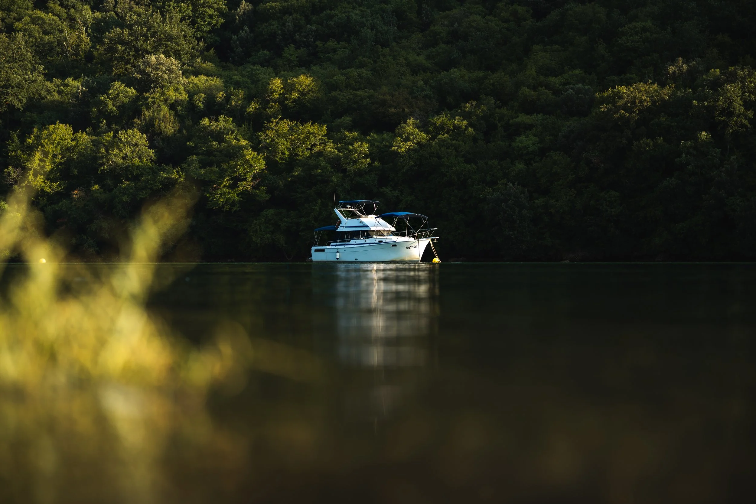 Motoryacht liegt ruhig in einem See vor einem bewaldeten Hügel, umgeben von grünen Bäumen, bei Sonnenuntergang.
