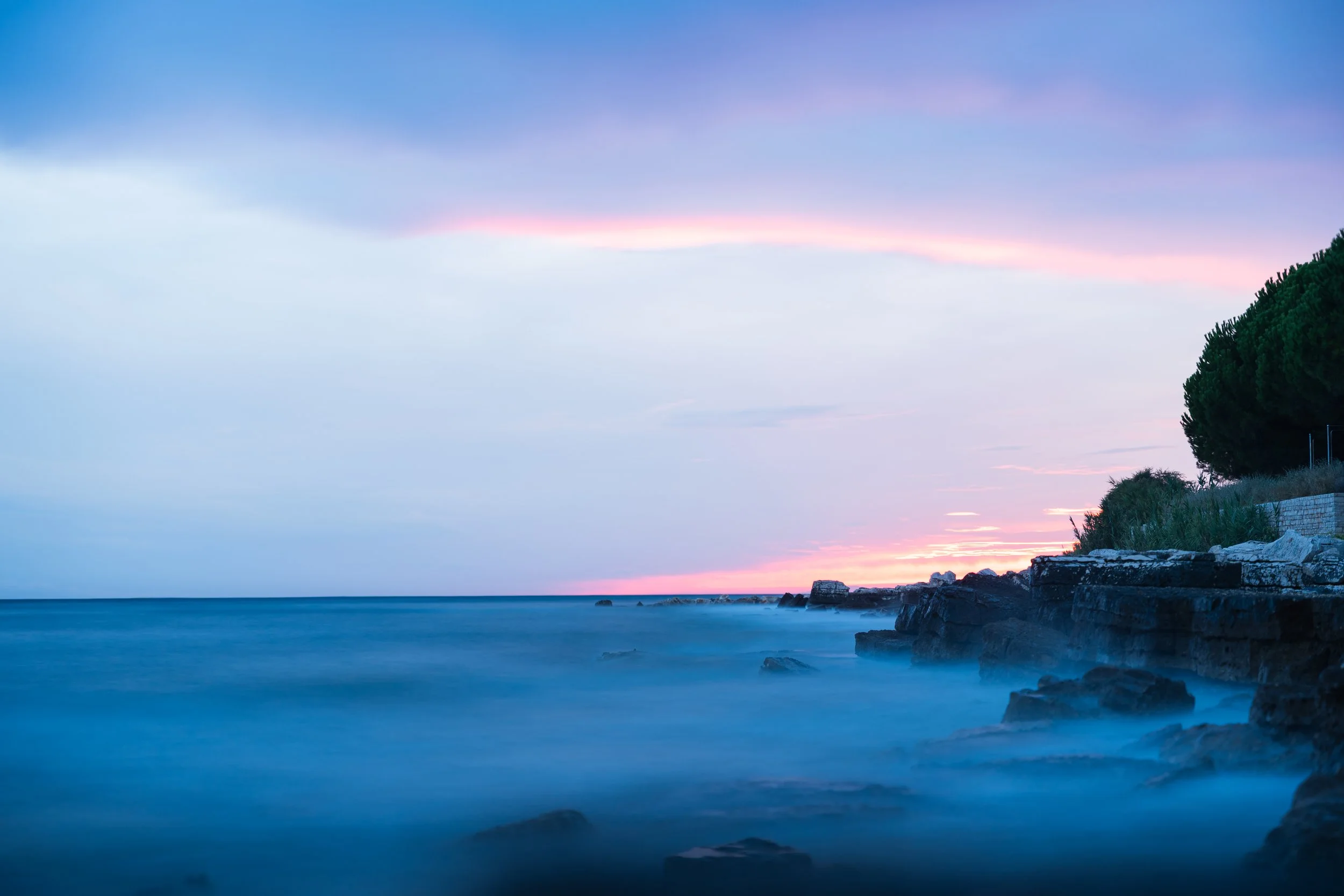 Ein ruhiger Küstenabschnitt bei Sonnenuntergang, mit ruhigem Wasser, Felsen und Bäumen am Ufer.
