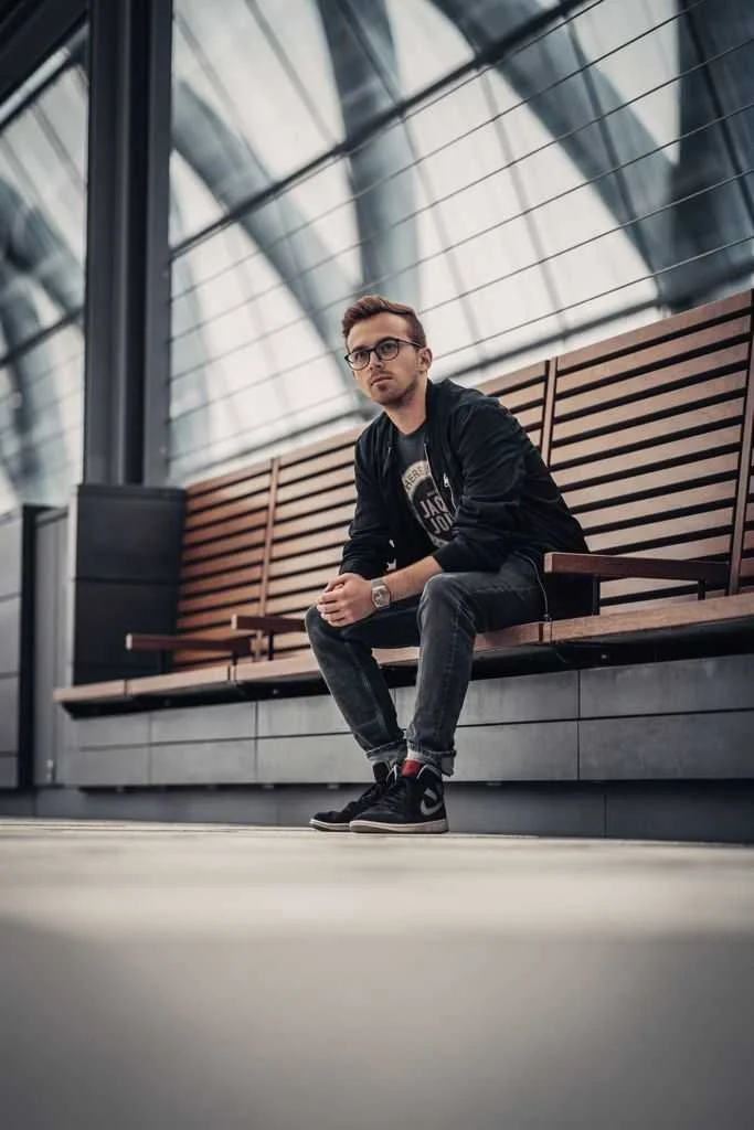 Ein junger Mann sitzt auf einer Bank in einer modernen, glasüberdachten Halle und schaut nach vorne.