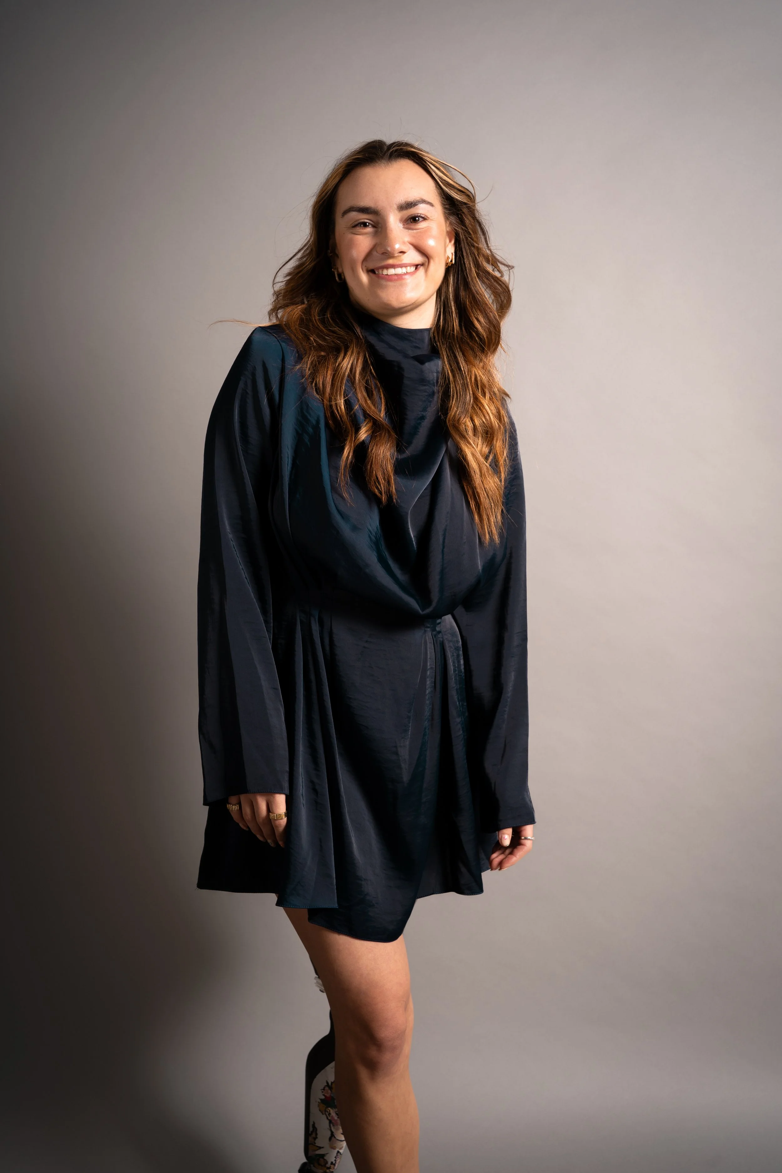 Eine lachende junge Frau mit braunen, gewellten Haaren, trägt einen schwarzen, glänzenden Kleidungsstil, steht vor einem neutralen grauen Hintergrund.