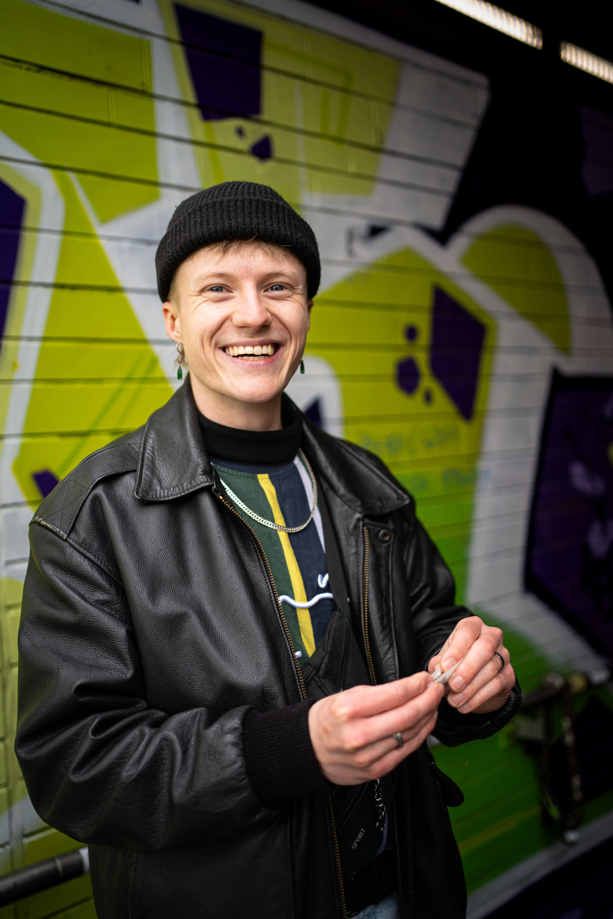 Junge Mann mit schwarzen Mütze, Lederjacke, im Hintergrund graffitiartige Wand