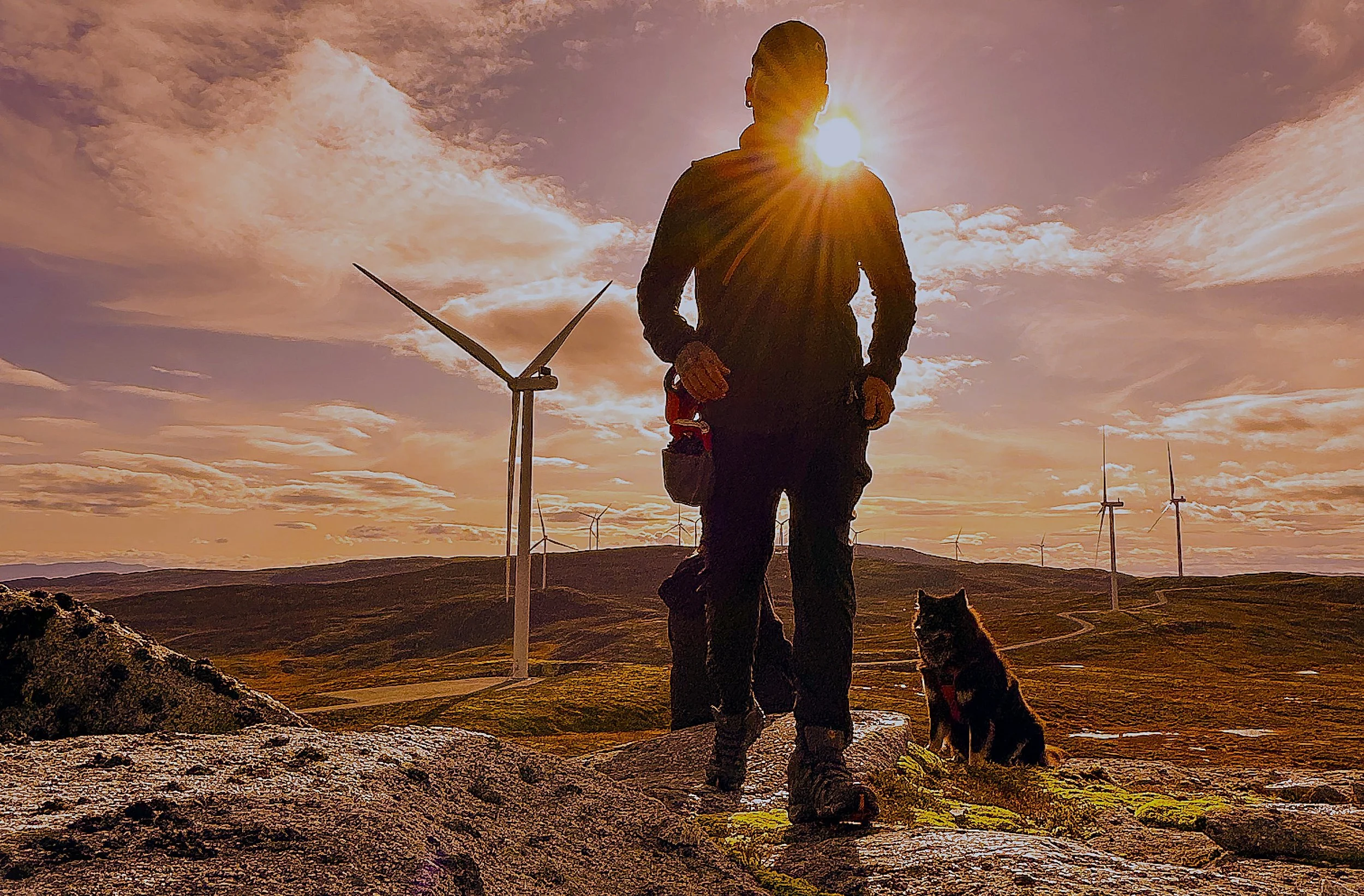 En person i utendørs antrekk med hund, solnedgang eller soloppgang, vindmøller i bakgrunnen, på en fjellandskap.