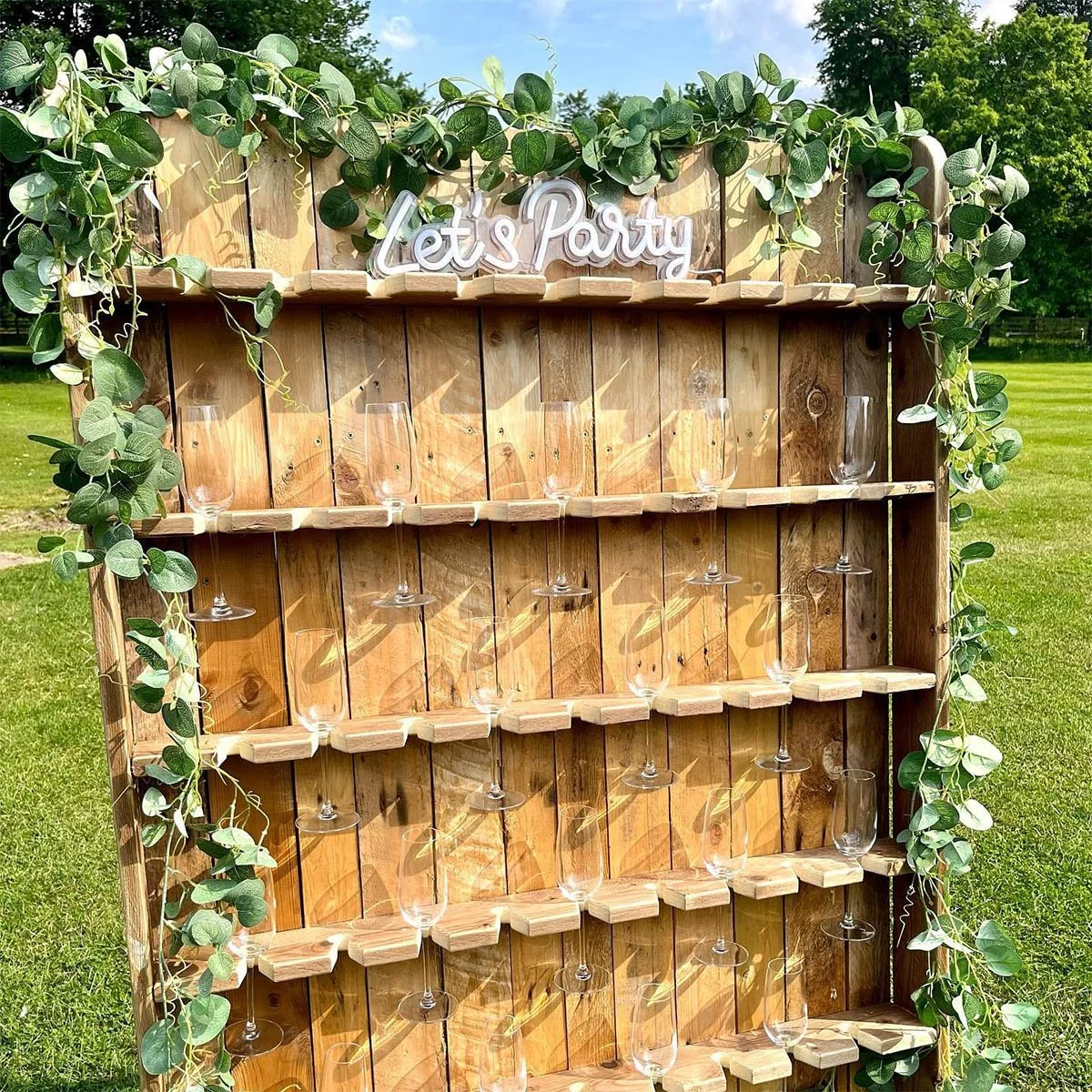 A wooden drink display board with greenery and a sign that says 'Let's Party' at the top. It has multiple shelves with empty wine glasses, set outdoors on a grassy area.