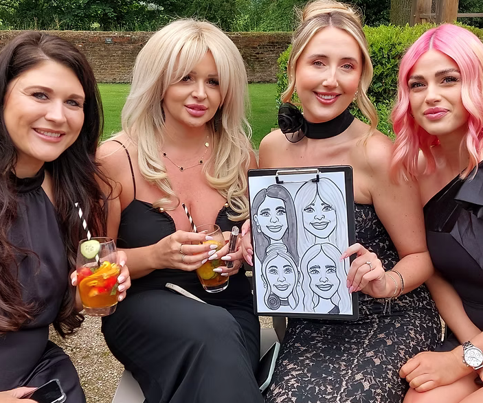 Four women sitting outdoors at a gathering, holding drinks, with a caricature drawing on a clipboard showing their portraits.