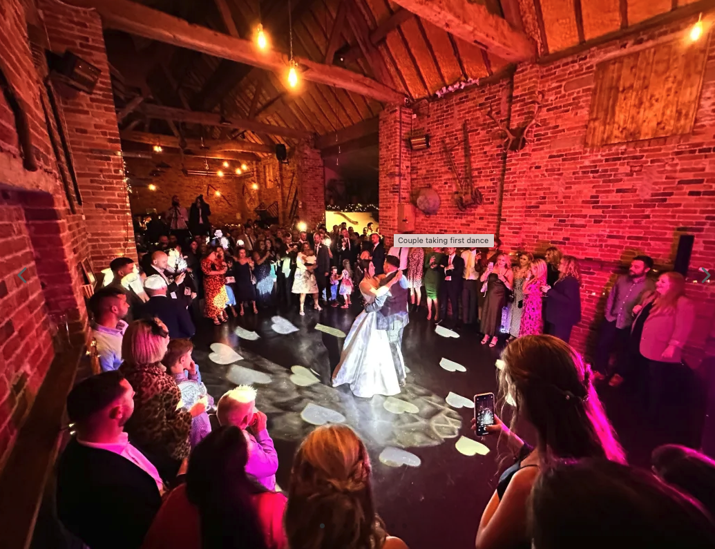 A couple is dancing in the center of a rustic venue with exposed brick walls, surrounded by guests watching and recording on their phones, during their first dance at a wedding reception.