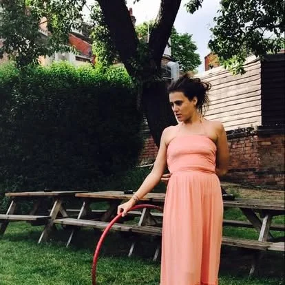Young woman in a peach-colored strapless dress standing in a yard with green grass, holding a red hula hoop, trees, and wooden benches in the background.