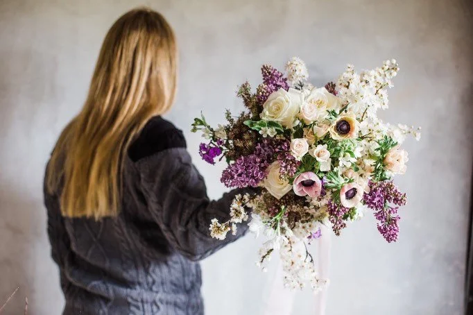 A woman with long blonde hair wearing a dark jacket holding a large bouquet of white, pink, and purple flowers against a plain background.