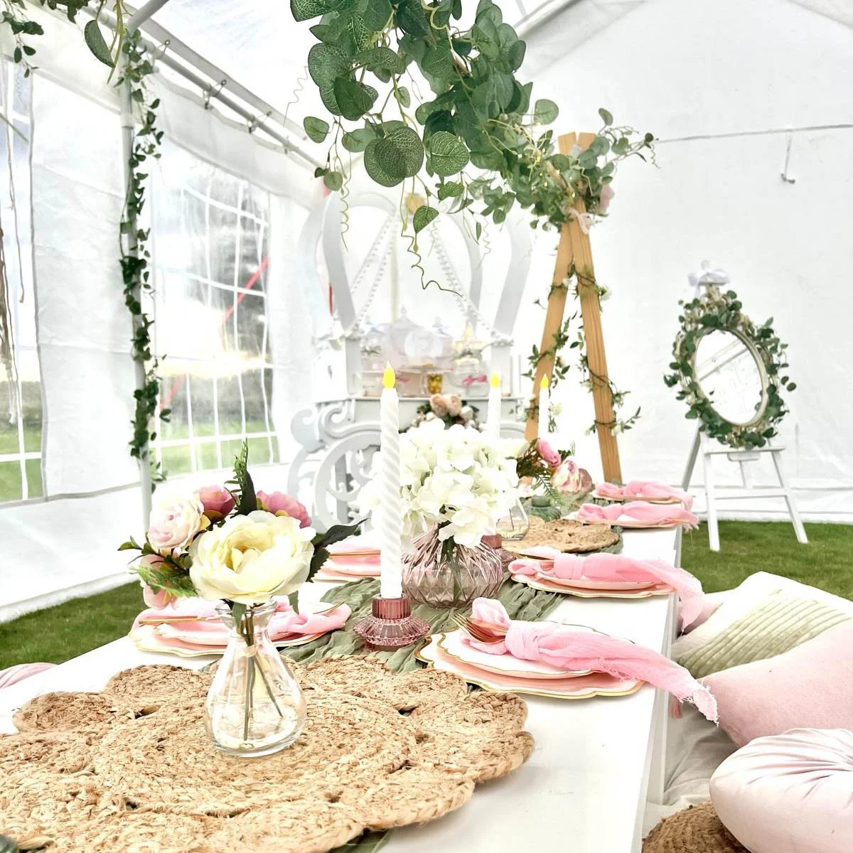 A decorated table setup inside a tent with floral centerpieces, pink napkins, and candles, likely for a wedding or celebration.
