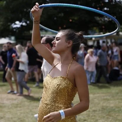 Young woman spinning a hula hoop at an outdoor event with a crowd in the background.