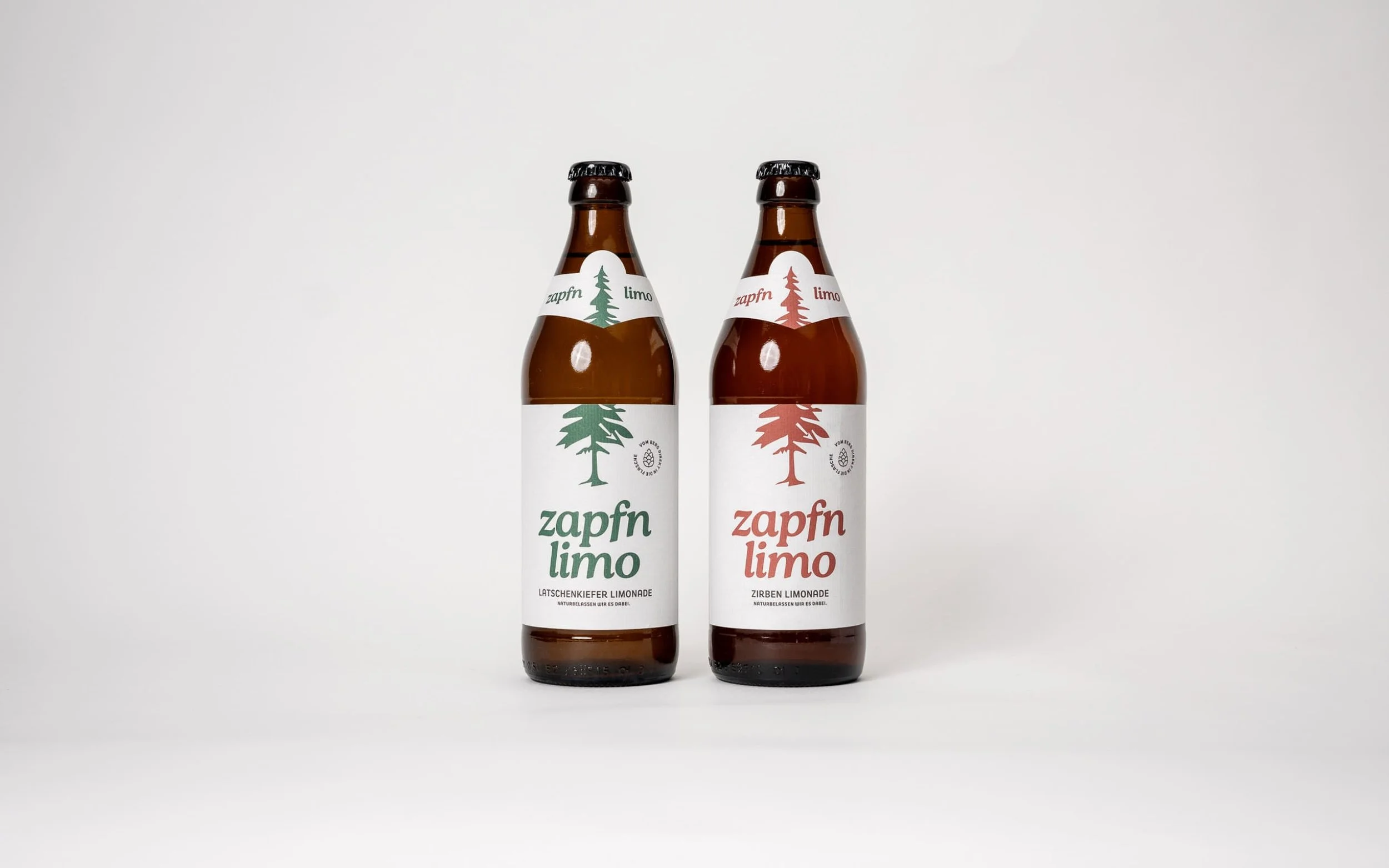Zwei Flaschen Zapfn Limo auf weißem Hintergrund, eine mit grünem Etikett "Latschenkiefer Limonade" und eine mit rotem Etikett "Zirben Limonade".