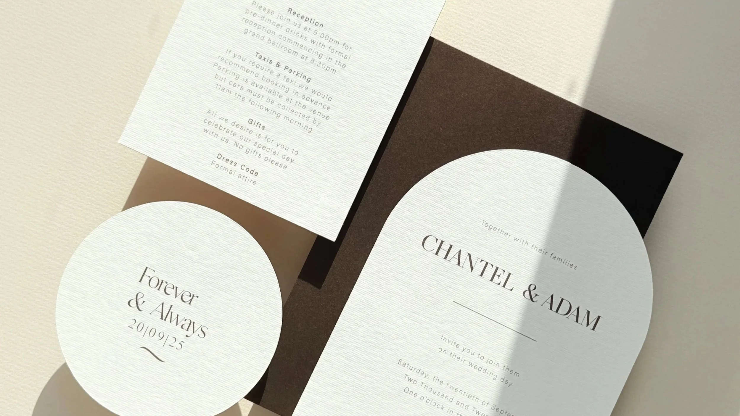 Wedding invitation card with the names Chantel and Adam, white circular tag with the words Forever & Always and the date 29/09/25, and a white paper with event details, placed on a dark and light backdrop.