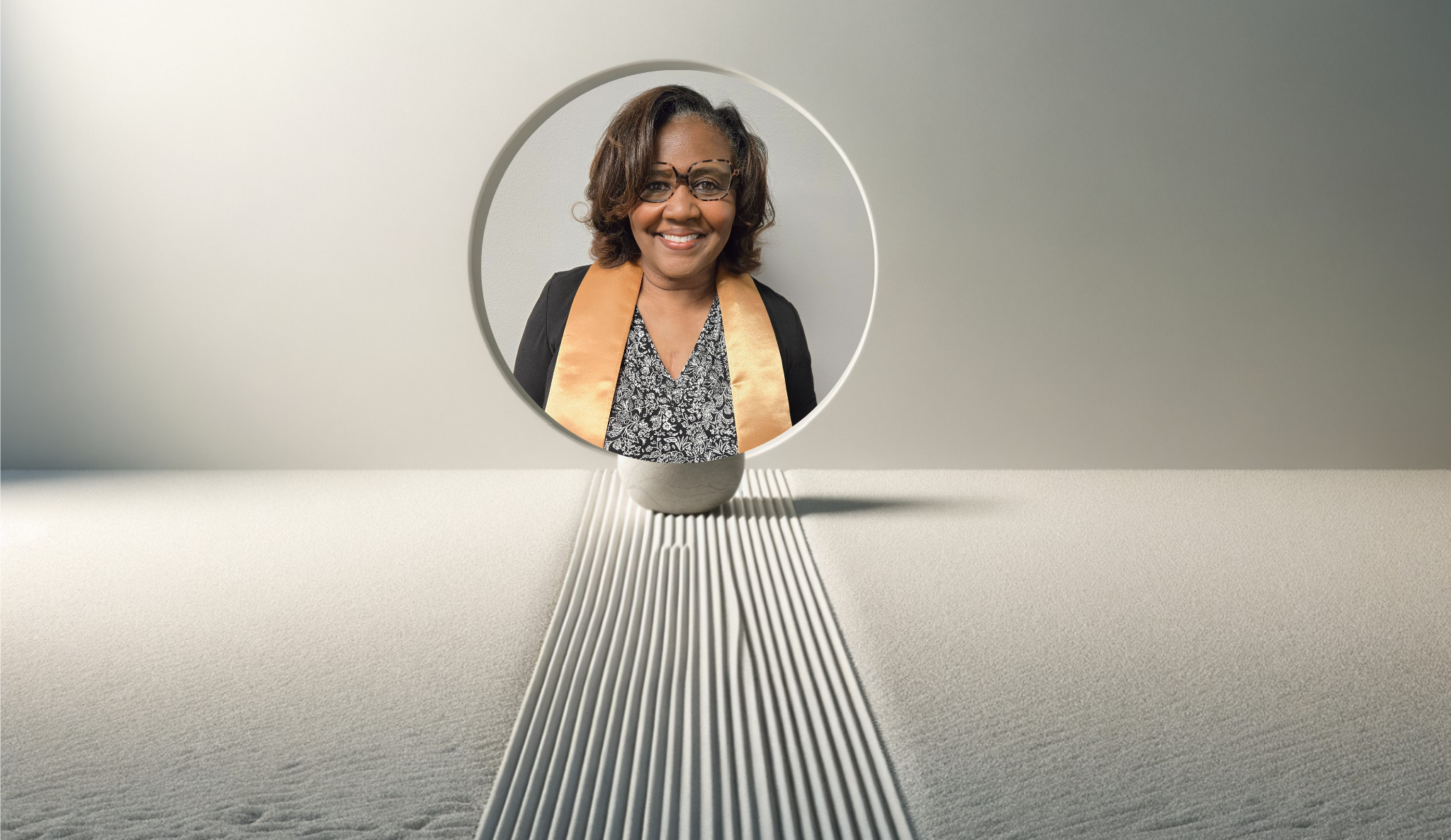 A woman with glasses, short hair, and a warm smile, dressed in a black blazer, patterned top, and a gold sash, reflected in a round mirror on a textured white surface.