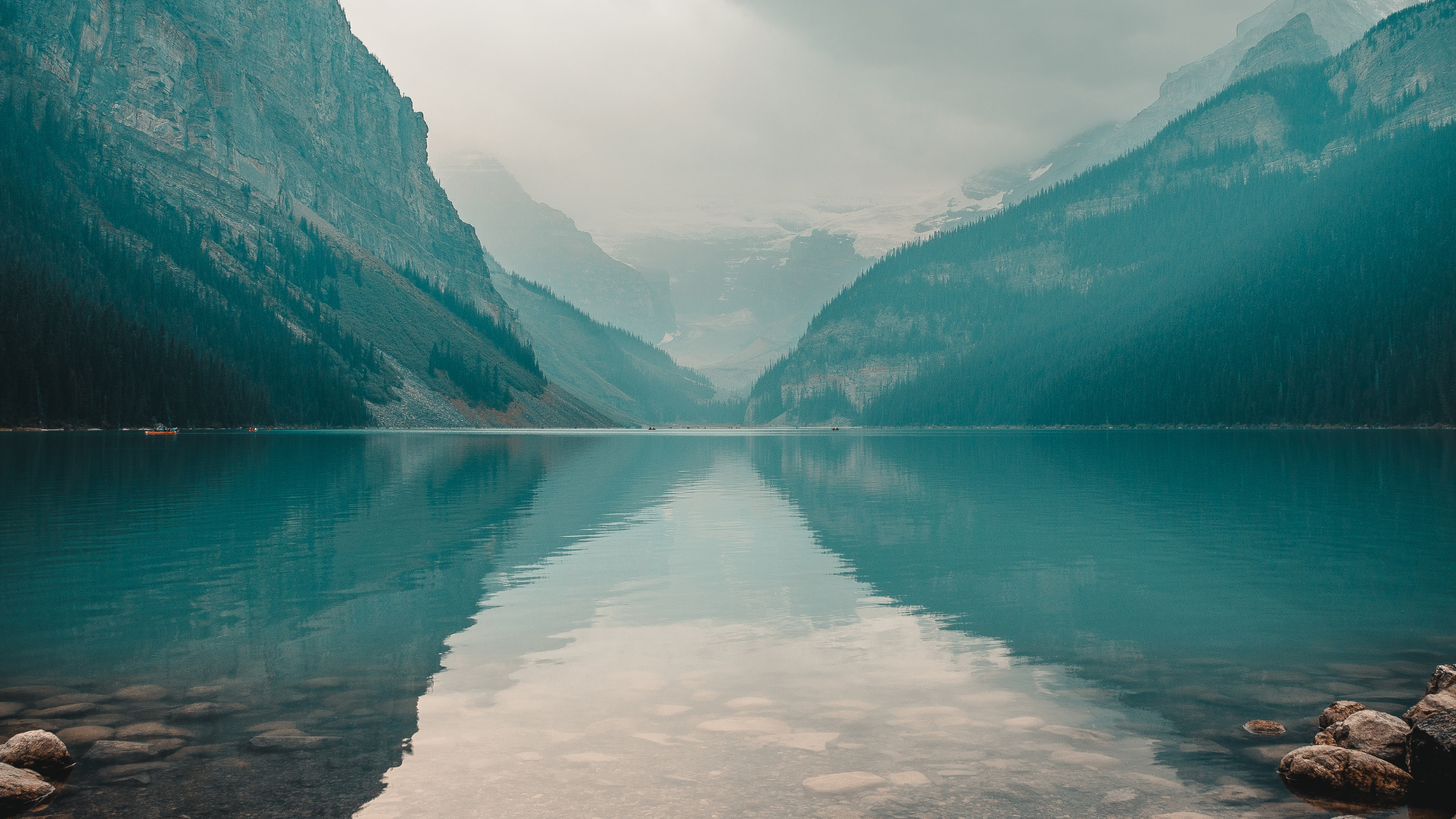 A calm mountain lake with turquoise waters reflecting the surrounding forested mountains and cloudy sky.