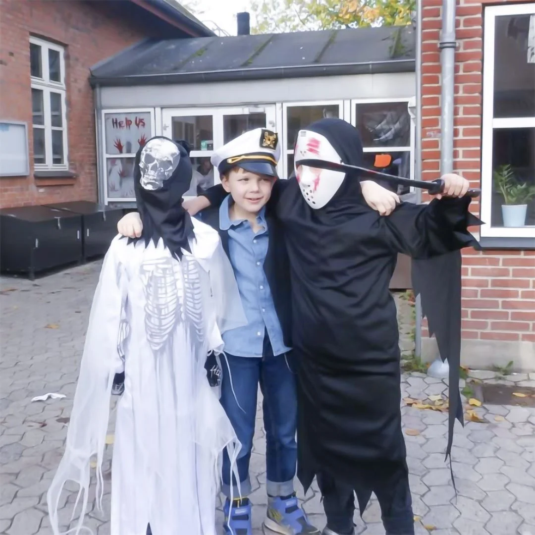 En dreng i politihat og blå skjorte med to personer i halloweenkostumer, en med skaldet maske og anden med sort kappe og blodige masker, der poserer sammen udendørs.