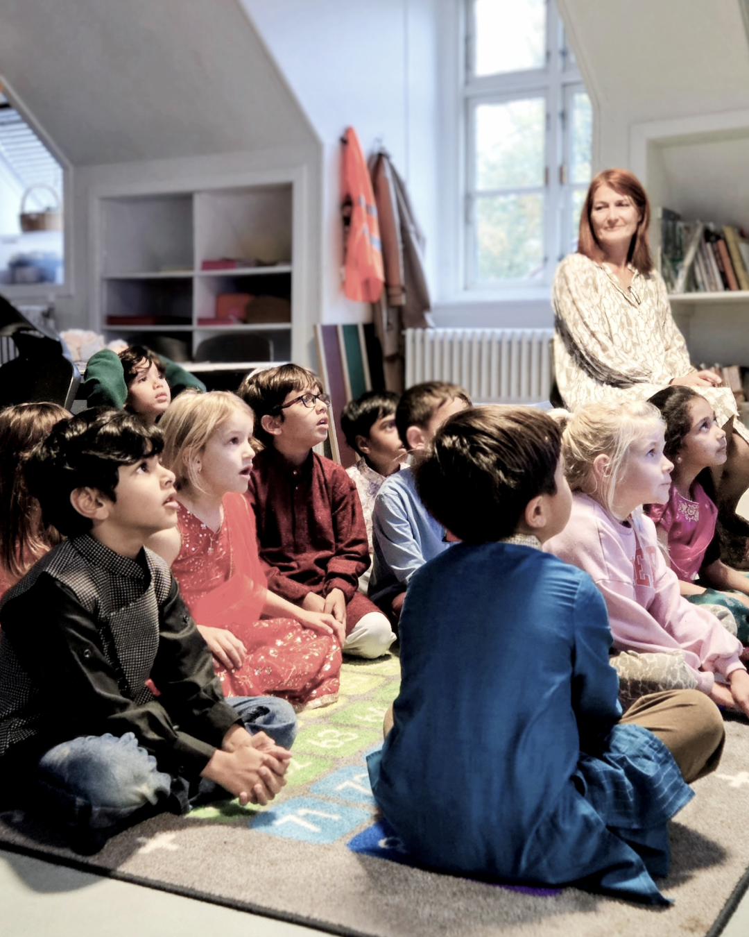 En voksen kvinde sidder sammen med en gruppe børn, der sidder på et tæppe i et klasseværelse og lytter opmærksomt.