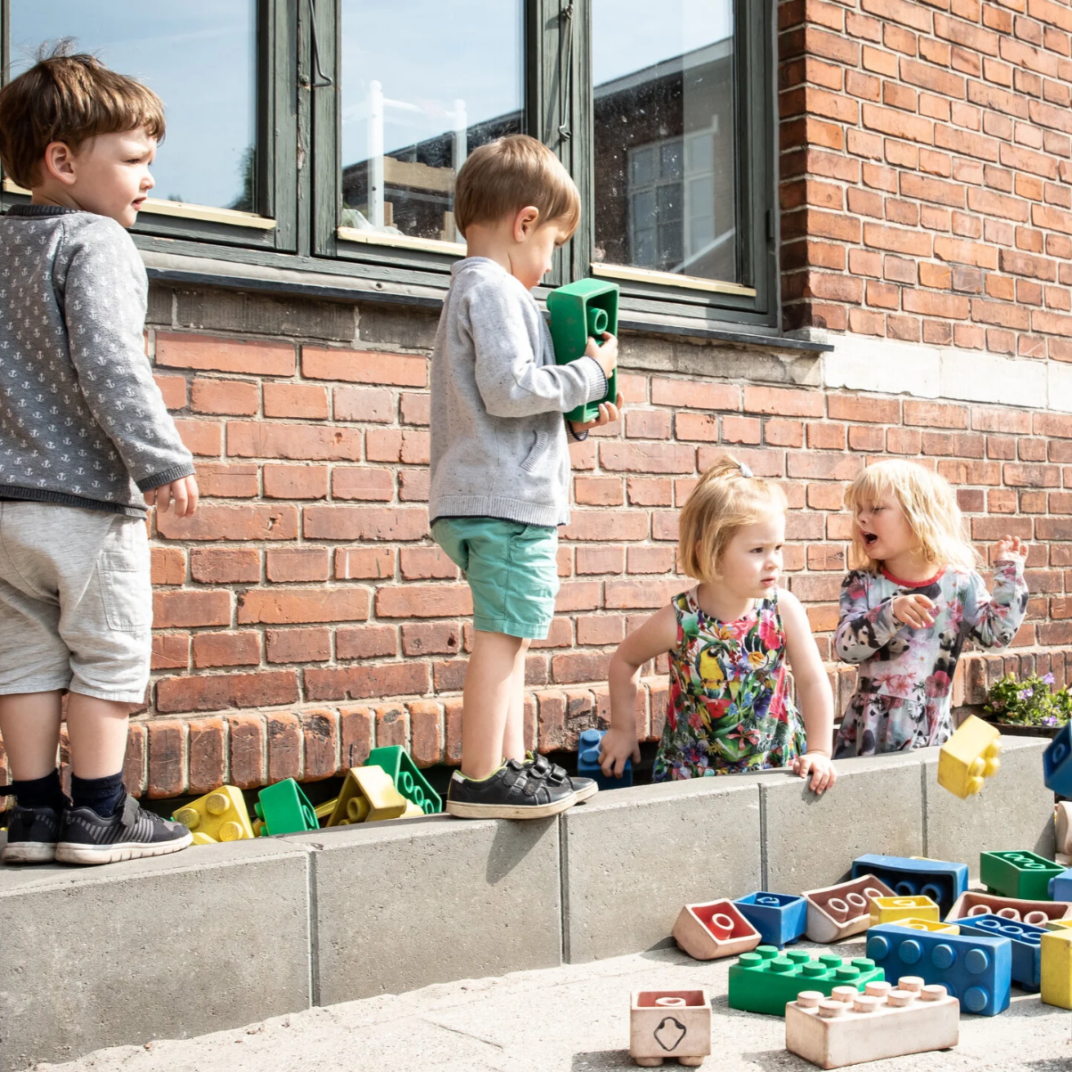 Fire børn leger med store farvede legoklodser udenfor ved en mur og et vindue.