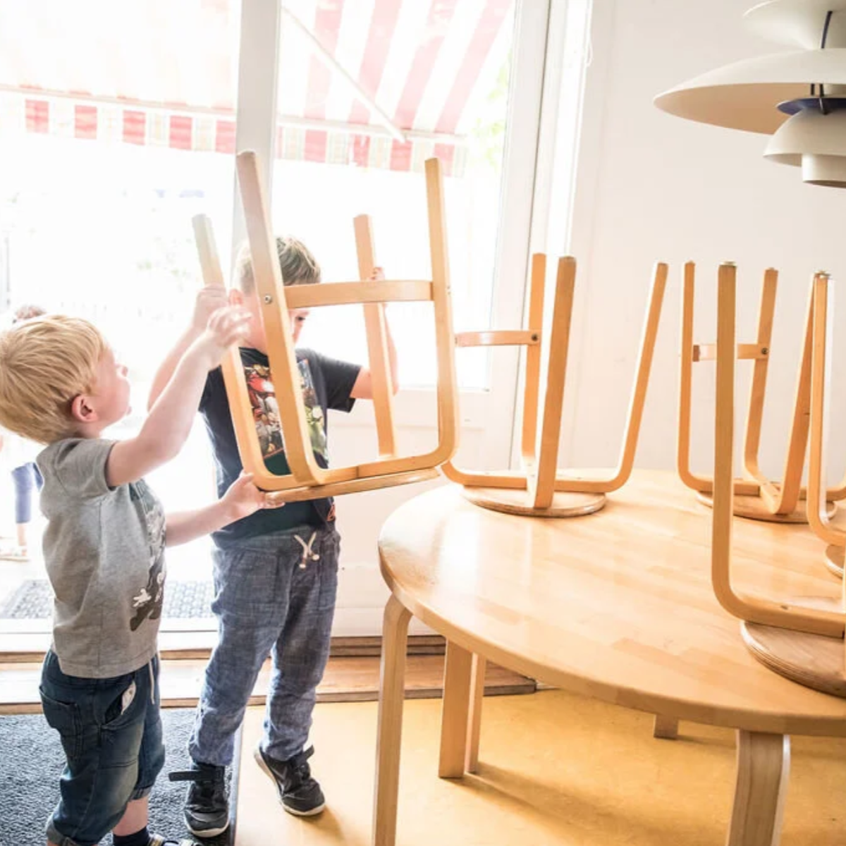 To børn løfter en trækantet træstol på et rundt bord, med flere ligestillede stole oppe på bordet.