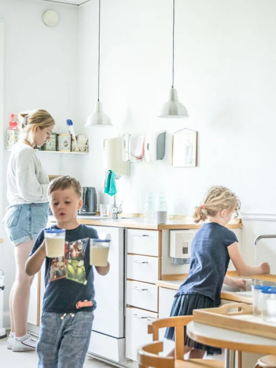 En kvinde og to børn i et køkken, hvor den ene dreng bærer to glas med væske, mens en pige vasker hænder ved vasken.