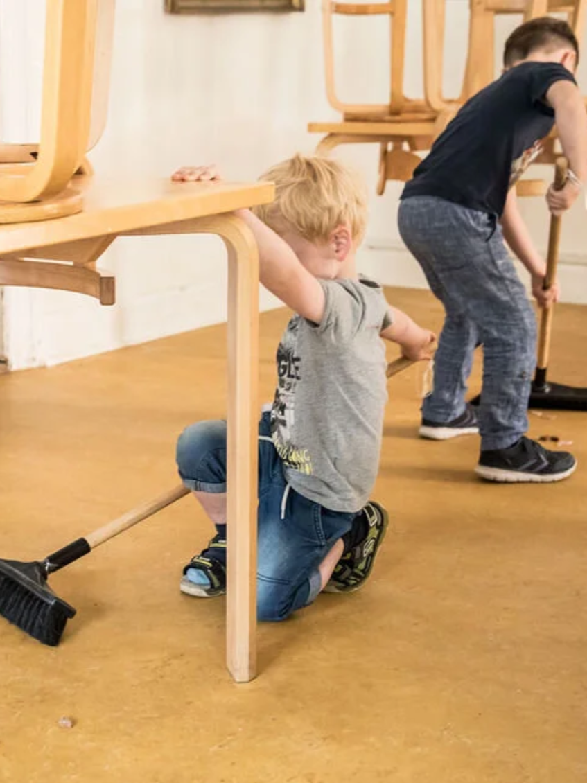 To børn leger under et bord, den ene knæler på gulvet og holder fast i bordet, den anden holder et spyd og står.