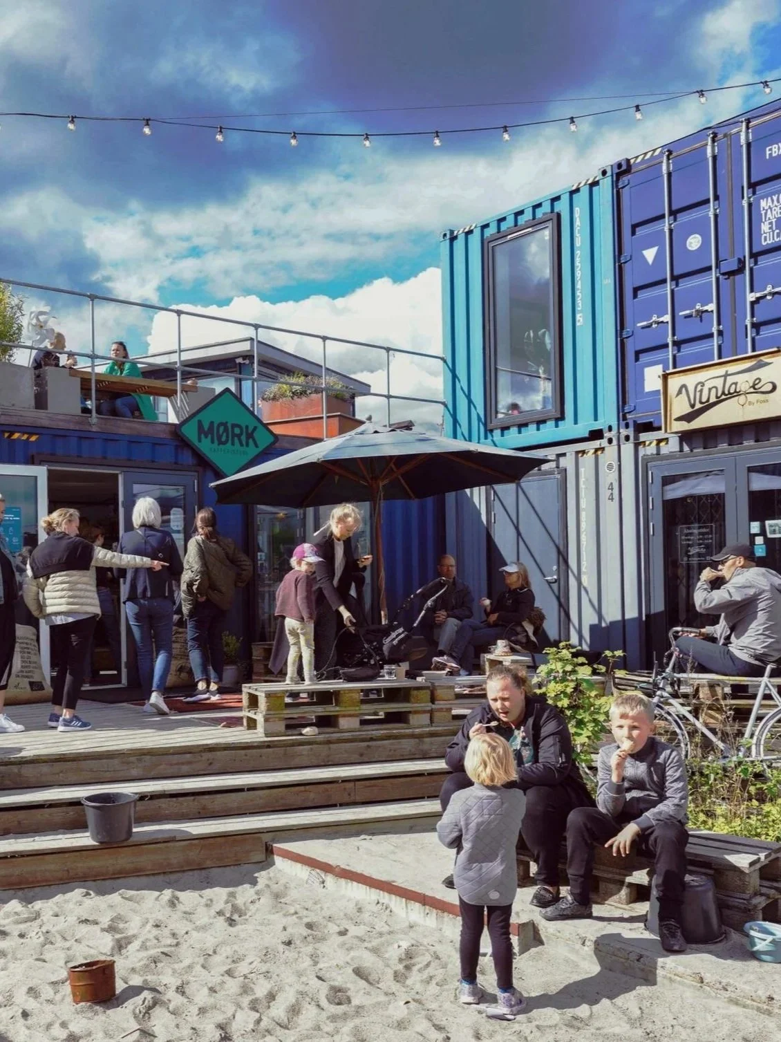 Gruppe mennesker nyder udendørs café med containerbygninger, står under paraply, børn leger i sandkasse, cykel og belysningskabler i blåt vejr.