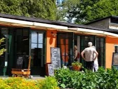 En person står foran en café eller restaurant med store vinduer og udendørs siddepladser, omgivet af grønne planter.