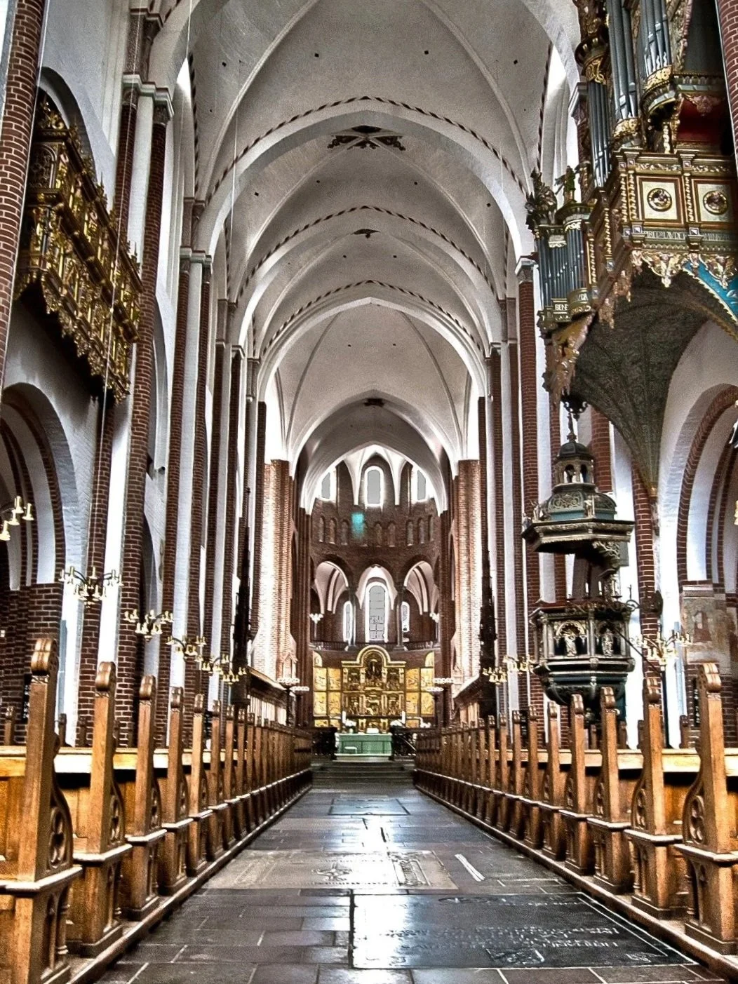 Indvendig af en gotisk kirke med høje hvide buede lofter, træbænkebænke langs siderne, et alter i den modsatte ende og en stor organ på højre side.