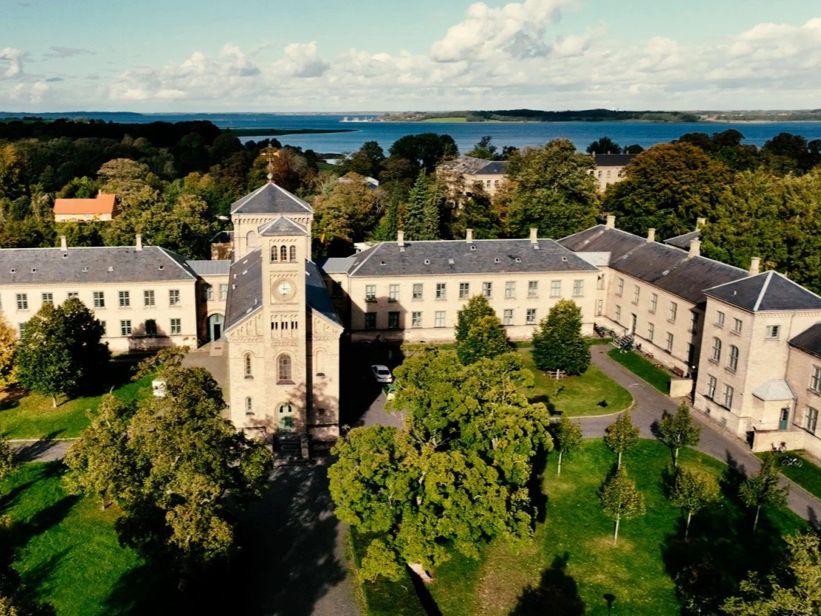 Billedet viser en ældre skolbygning med en tårn på midten, omgivet af grønne træer og en sti. Der er en krop af vand i baggrunden.