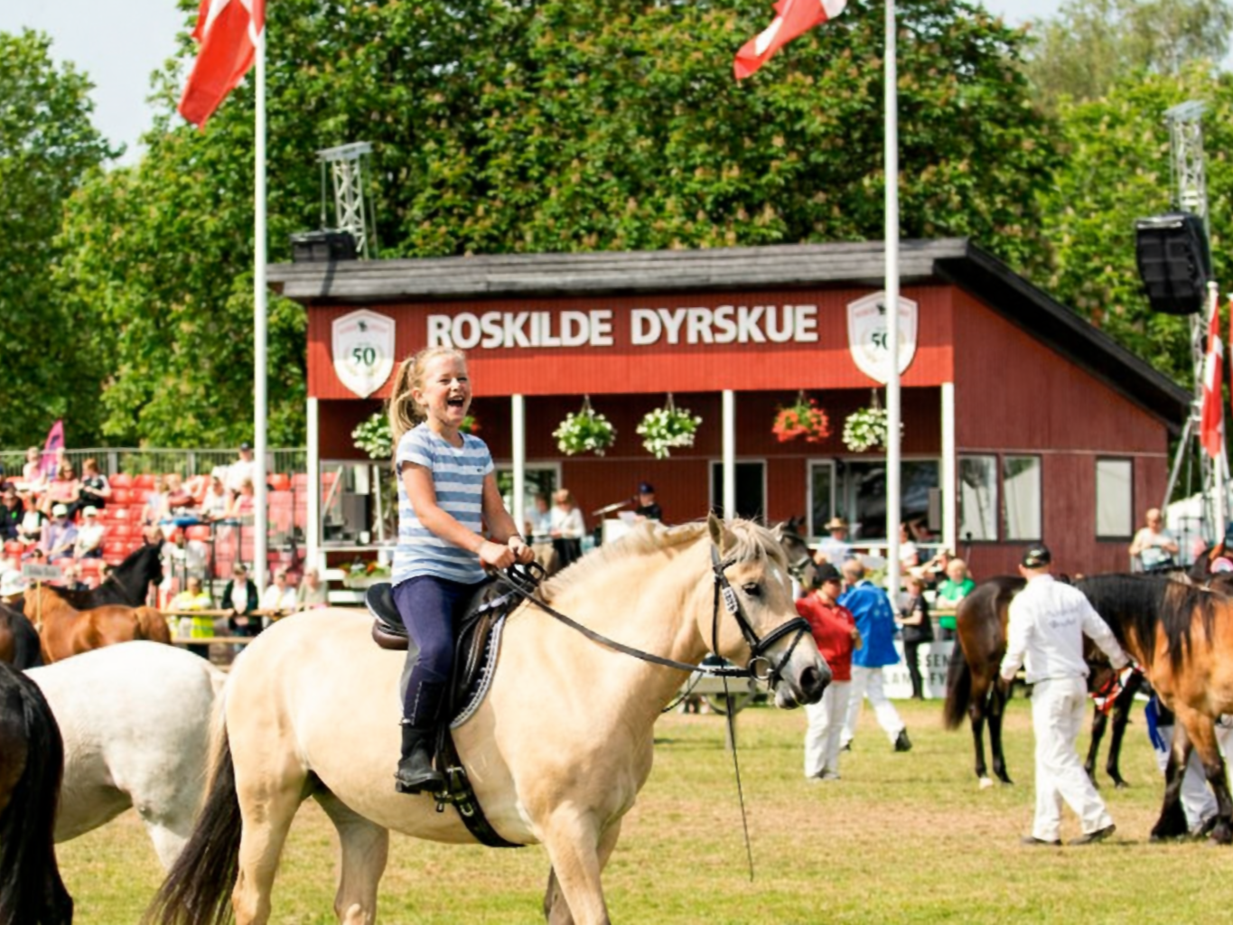 En ung pige rider på en hest under en festival eller et event med mange mennesker og andre heste i baggrunden.