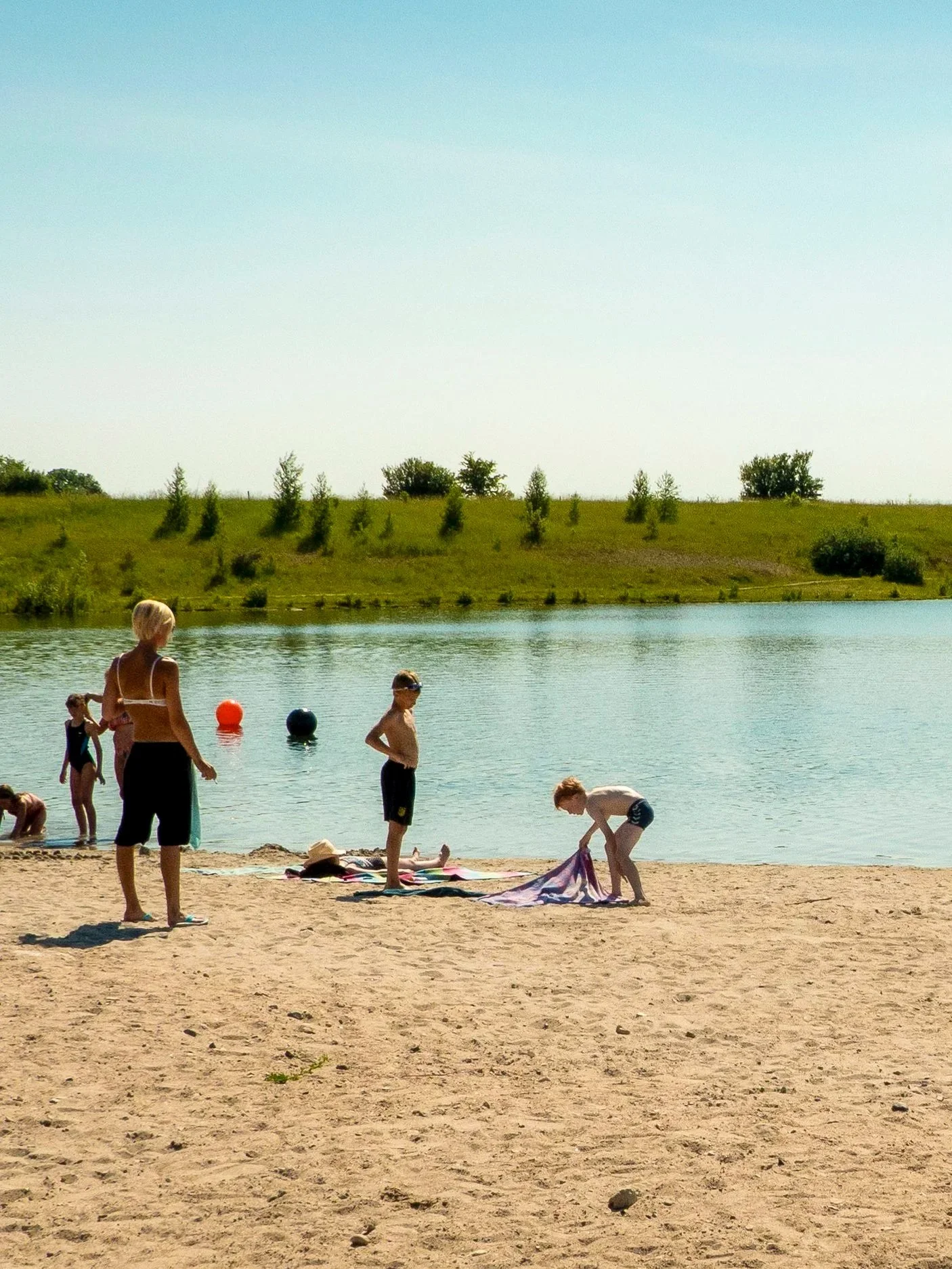 Børn leger på en strand ved en sø, nogle er i vandet, nogle leger med ting på sandet, med en grøn bakke i baggrunden under en klar blå himmel.