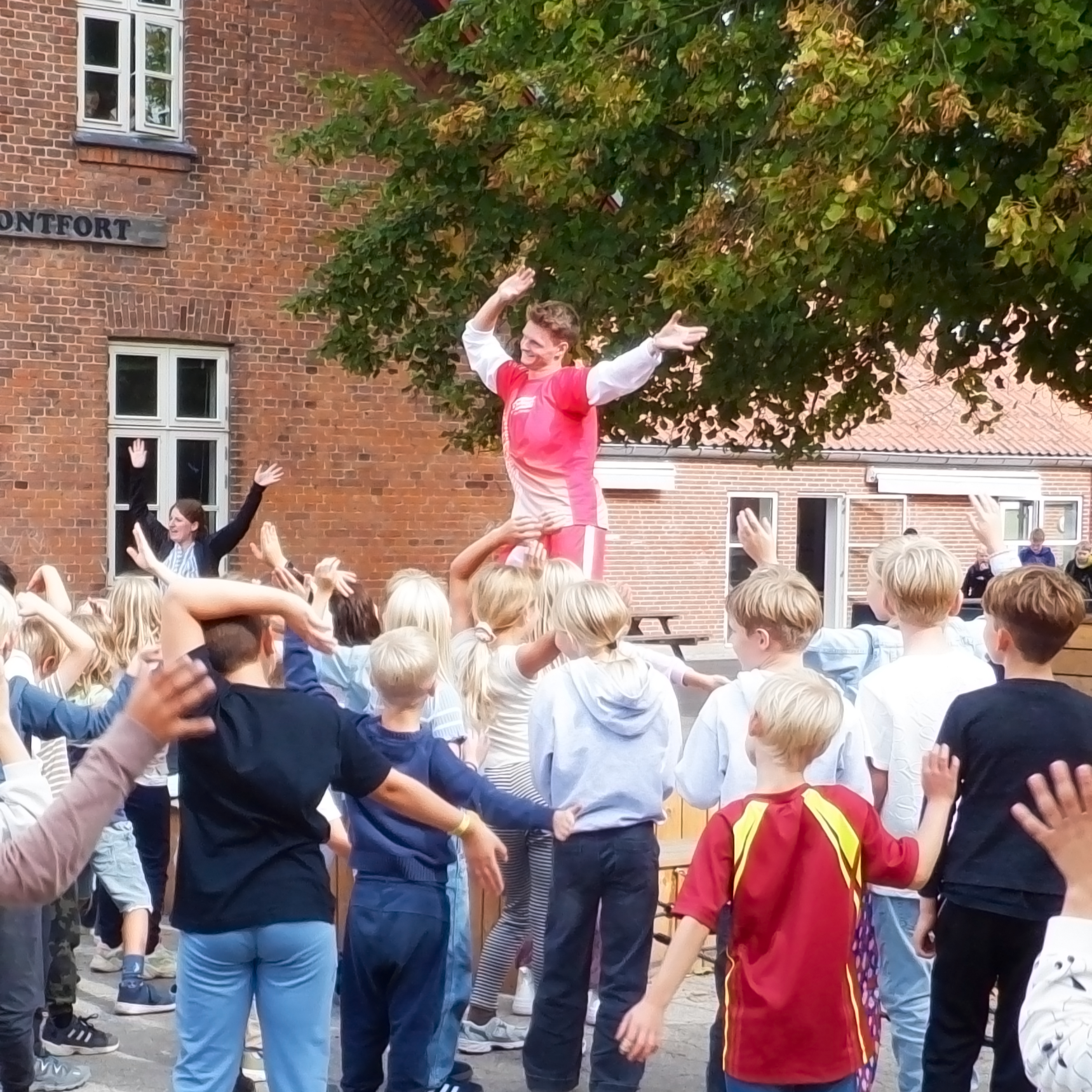 En gruppe børn danser udenfor, med en performer på skuldrene, foran et rødt murstenshus og et træ.