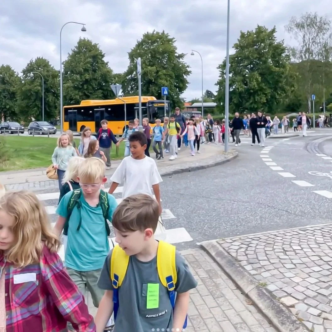 Børn krydser gaden på skoleudflugt, med bus i baggrunden og grønne træer i baggrunden.