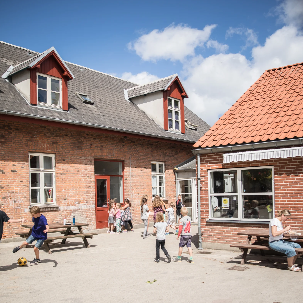 Børn leger udendørs foran en rød murstensbygning med flere vinduer og tage under en blå himmel med få skyer.