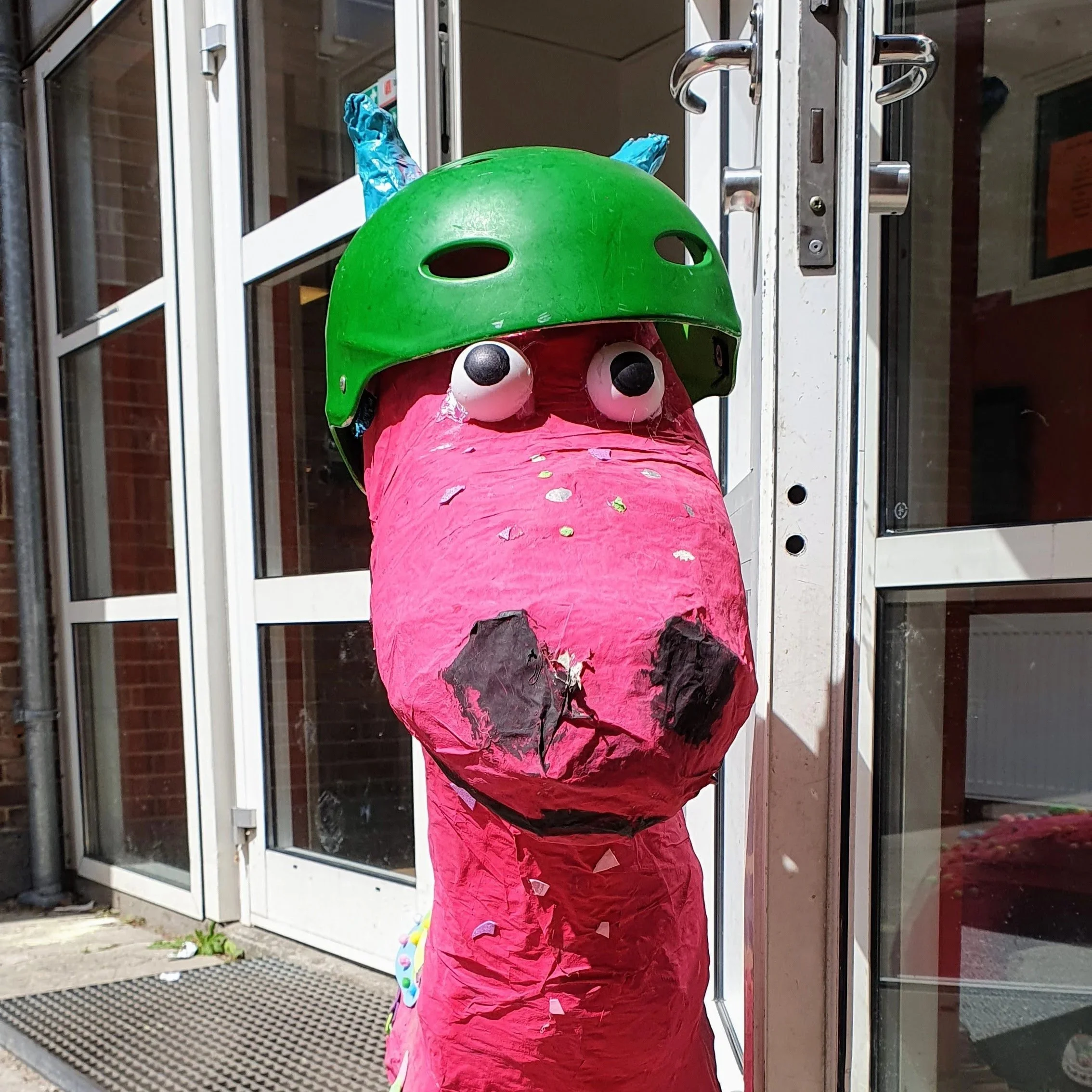 En farverig artistisk næsefigur lavet af papmaché med en grøn cykelhjelm som hat, placeret foran en glasdør.