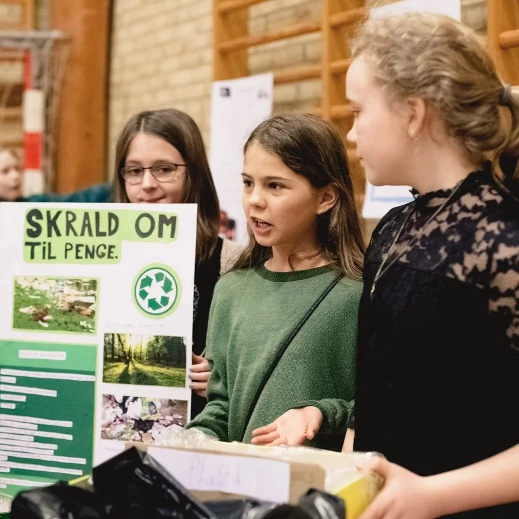 Tre unge piger taler om affald og genbrug ved en informationsstand med plakater om genbrug.