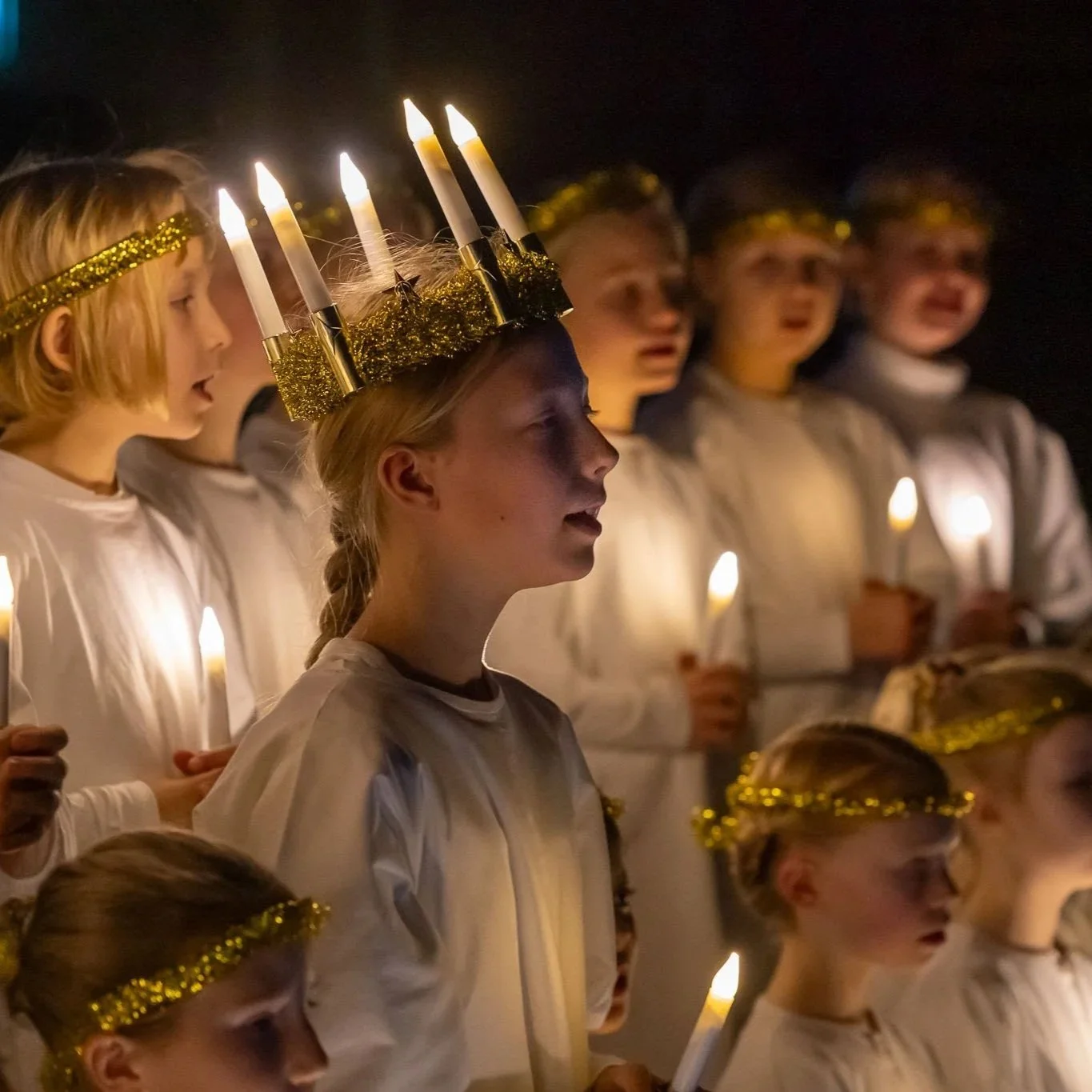 En gruppe børn iført hvide kostumer, nogle med guldkranse og en pige med en gylden krone, holder lysende fakler under en ceremoni om aftenen.