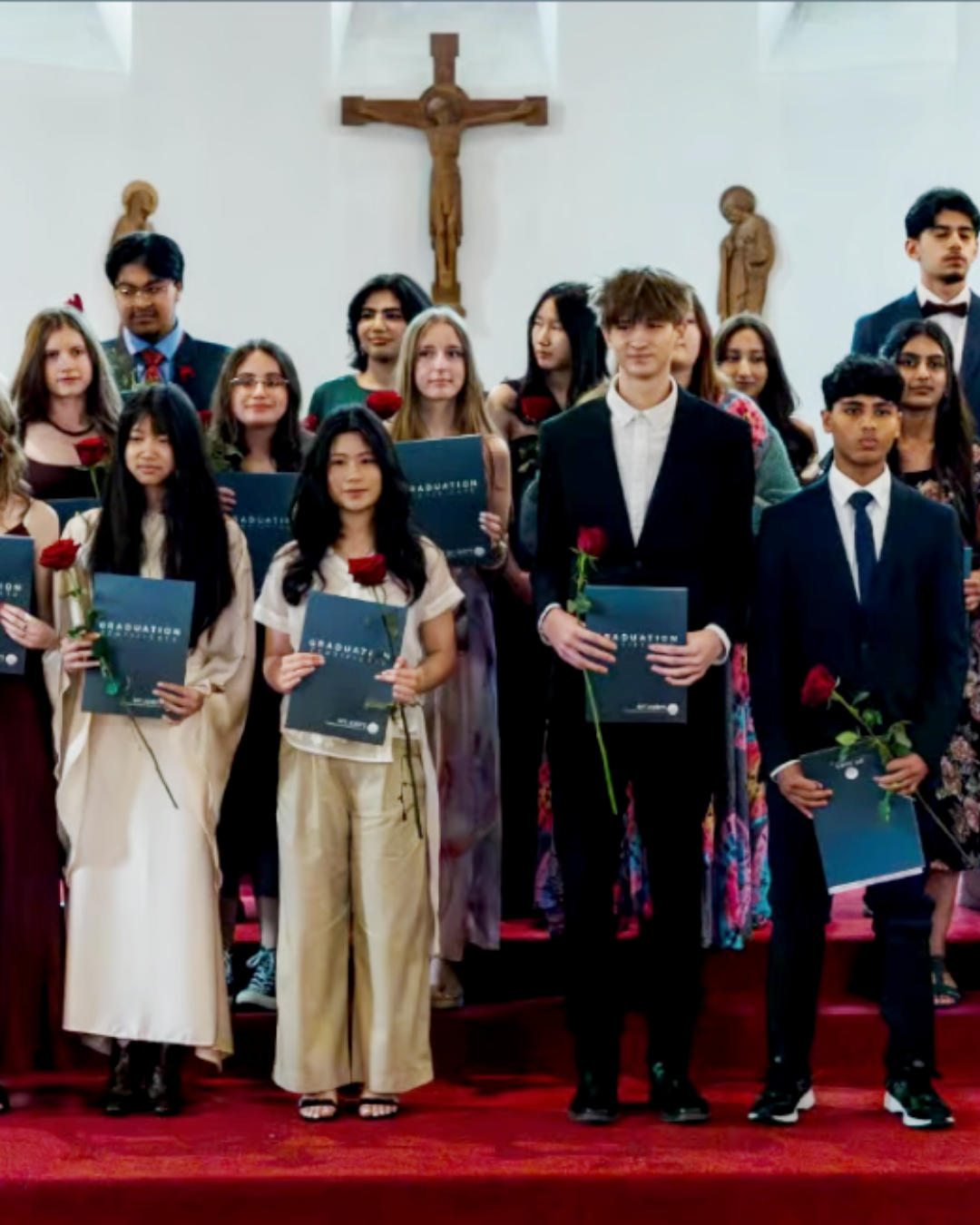 Gruppe af unge med diplomer og roser til en gymnasiestartceremoni i en kirke med kors og religiøse statuer i baggrunden.