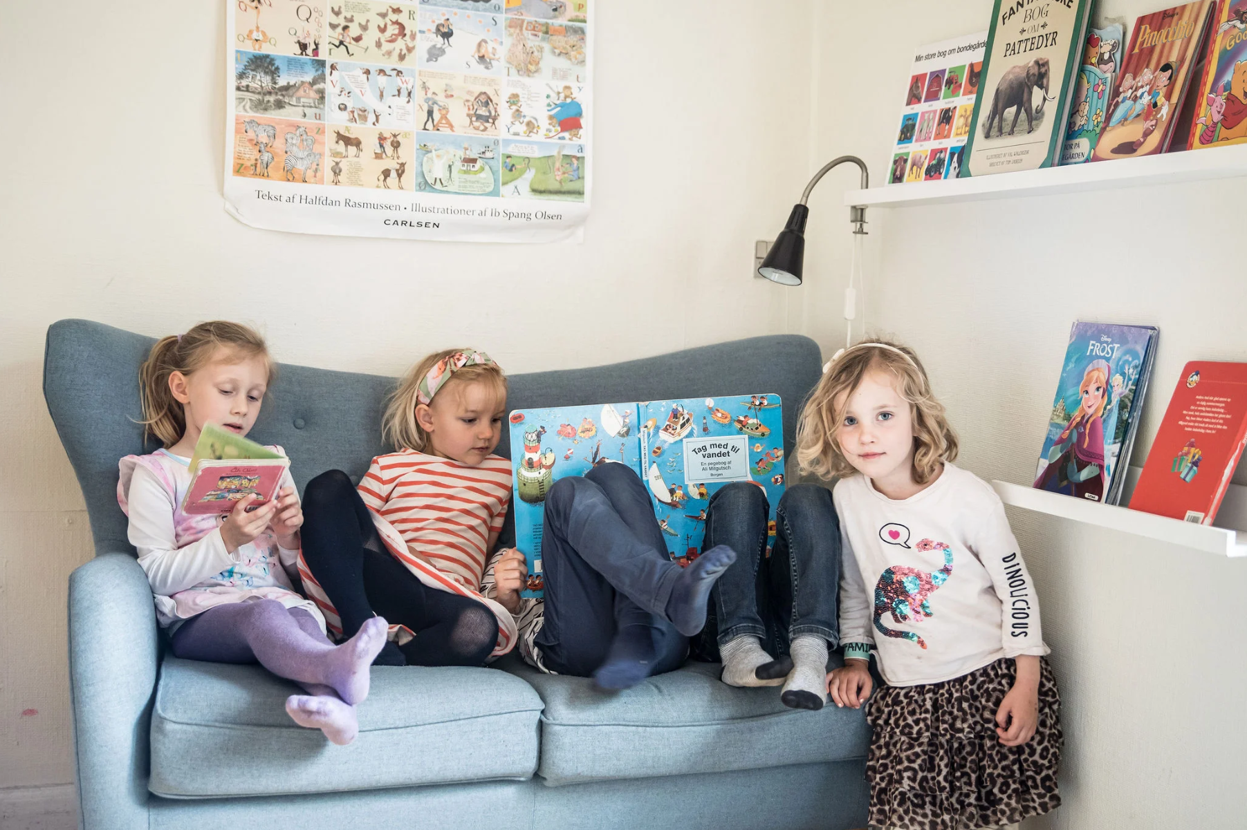 Fire unge børn sidder på en sofa og læser bøger i et hyggeligt rum med bogreoler og posters på væggen.