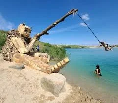 En skulptur af en bjørn, der fisker med en stang på en strand ved en sø